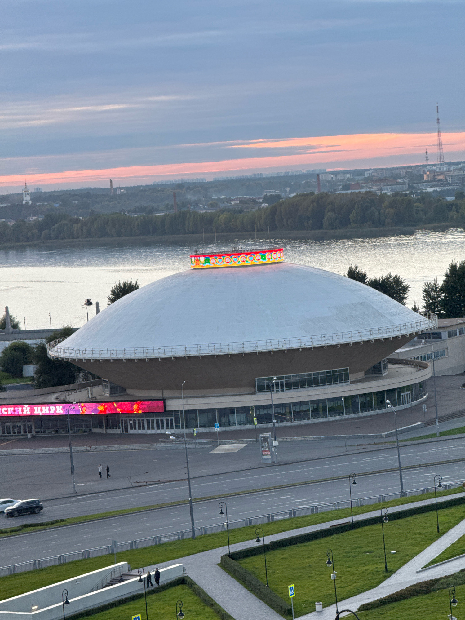 The image shows a wide, low-angle exterior shot of the Kazan Circus building in Kazan, Russia, during dusk. The circus building is characterized by its distinctive, saucer-shaped roof, which is white and bordered by a decorative illuminated ring of colorful lights around its upper edge. Below the main dome, a multi-story structure with large glass windows is visible. A vibrant LED display screen is mounted on the lower facade of the building, showing colorful graphics and the text "КРАСКИ ЦИРКА" (meaning "Paints of the Circus") and "СКИЙ ЦИРК" (likely part of the Kazan Circus name).
In the foreground, a multi-lane road runs in front of the circus. Several cars are present on the road, and two individuals are walking on the sidewalk in front of the circus building. Beyond the road, a grassy area with decorative lighting and pathways is visible, suggesting a park or public space. In the background, a wide river reflects the muted light of the sky, and beyond the river, a cityscape with trees, buildings, and a tall radio tower can be seen against a soft, hazy sky with streaks of pink and orange indicating the setting sun. The overall atmosphere is serene and somewhat subdued, typical of twilight in an urban environment.