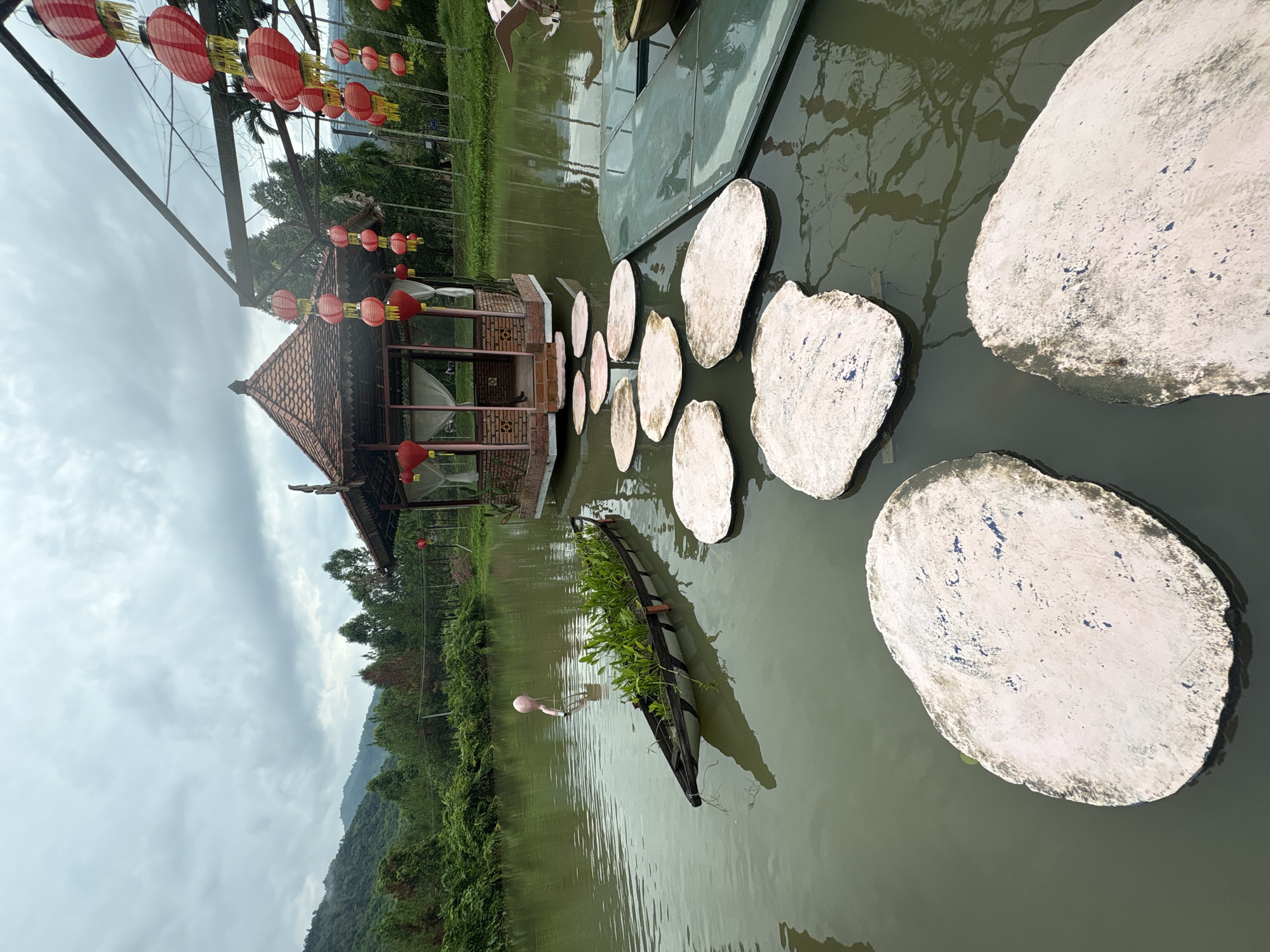 The image captures a serene scene in Khanh Vinh, Vietnam, featuring a small brick gazebo adorned with red lanterns, set within a pond. A series of stepping stones lead across the water towards the gazebo. A long, narrow boat filled with plants floats nearby. The surrounding environment includes lush greenery and a backdrop of mountains. Overcast skies suggest a cloudy day.