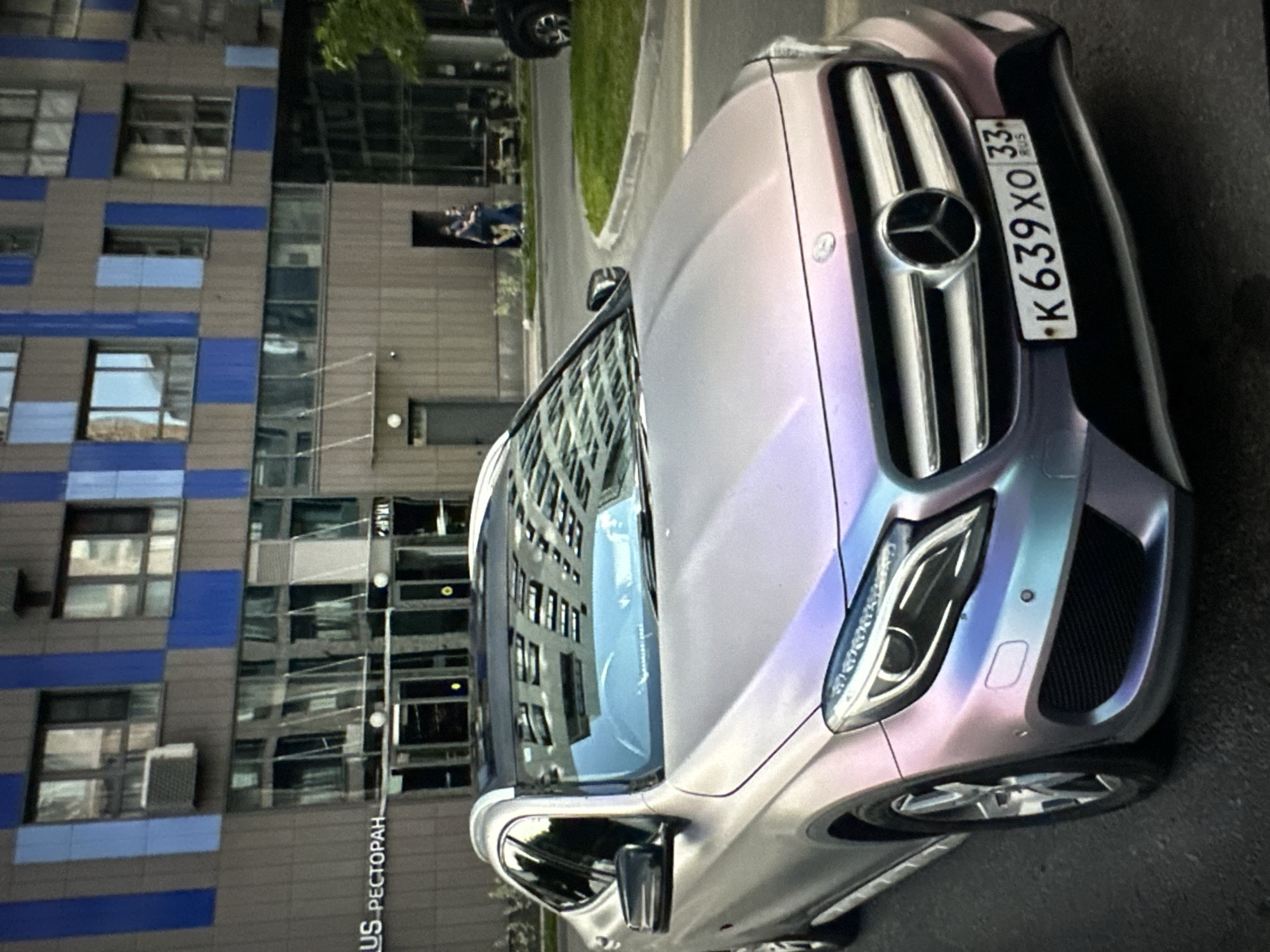 A pearlescent silver Mercedes-Benz GLA-Class (X156) is parked on a street in front of a modern building with blue and brown paneling. The car's license plate is visible and reads "K639XO 33 RUS". The building features large windows that reflect the surrounding environment. In the background, two people are standing near an entrance to the building. A patch of grass is visible to the right of the car, bordering the street. The lighting suggests it is daytime.