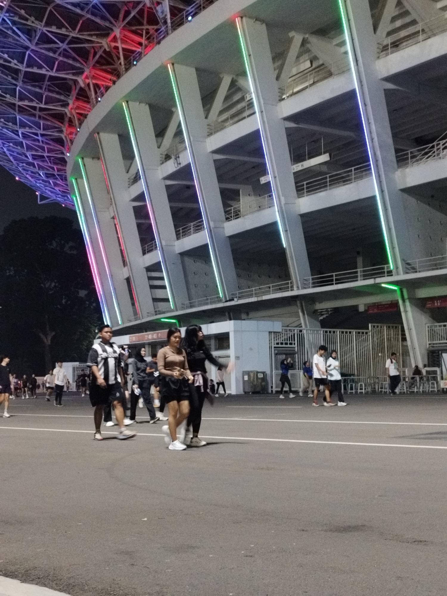 In Central Jakarta, Indonesia, an image captures a group of people walking outside a large stadium at night. The stadium features a modern, geometric structure illuminated by colorful LED lights along its exterior. The lighting displays a gradient of red, pink, green, and blue, creating a vibrant atmosphere. People, dressed casually in t-shirts, shorts, and athletic wear, are seen walking on a paved area in front of the stadium. Some are walking individually, while others are in pairs or small groups. One individual is seen on a skateboard. The background includes trees and the upper levels of the stadium structure with railings and support beams visible. The overall scene suggests a public event or gathering at the stadium.