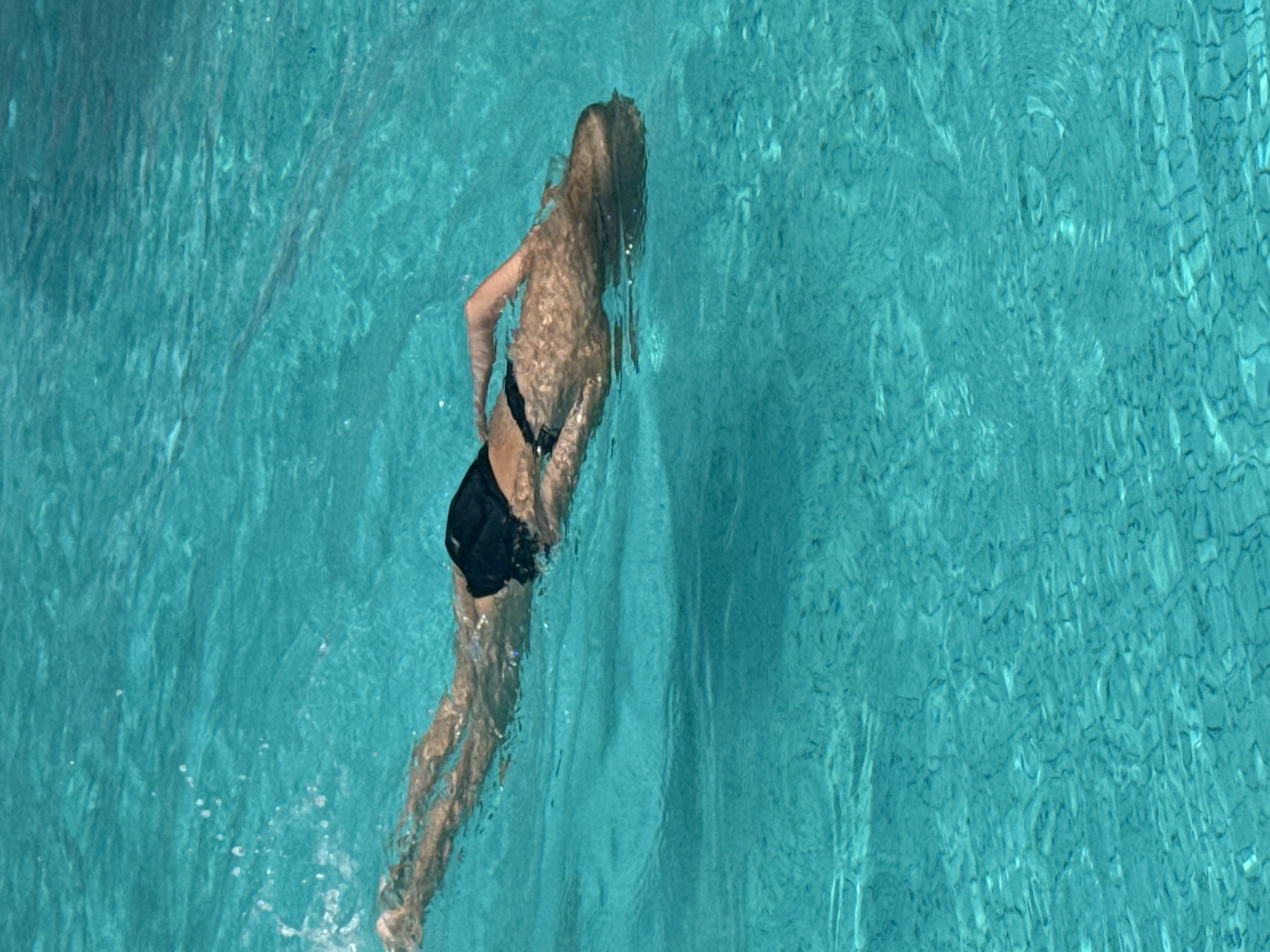 A person swims underwater in a pool with bright blue water. The person is wearing black swim trunks. Their body is mostly submerged, with only their back and head visible at the surface. The water is disturbed by their movement, creating ripples and splashes. The bottom of the pool is visible beneath the clear water, showing a mosaic tile pattern.