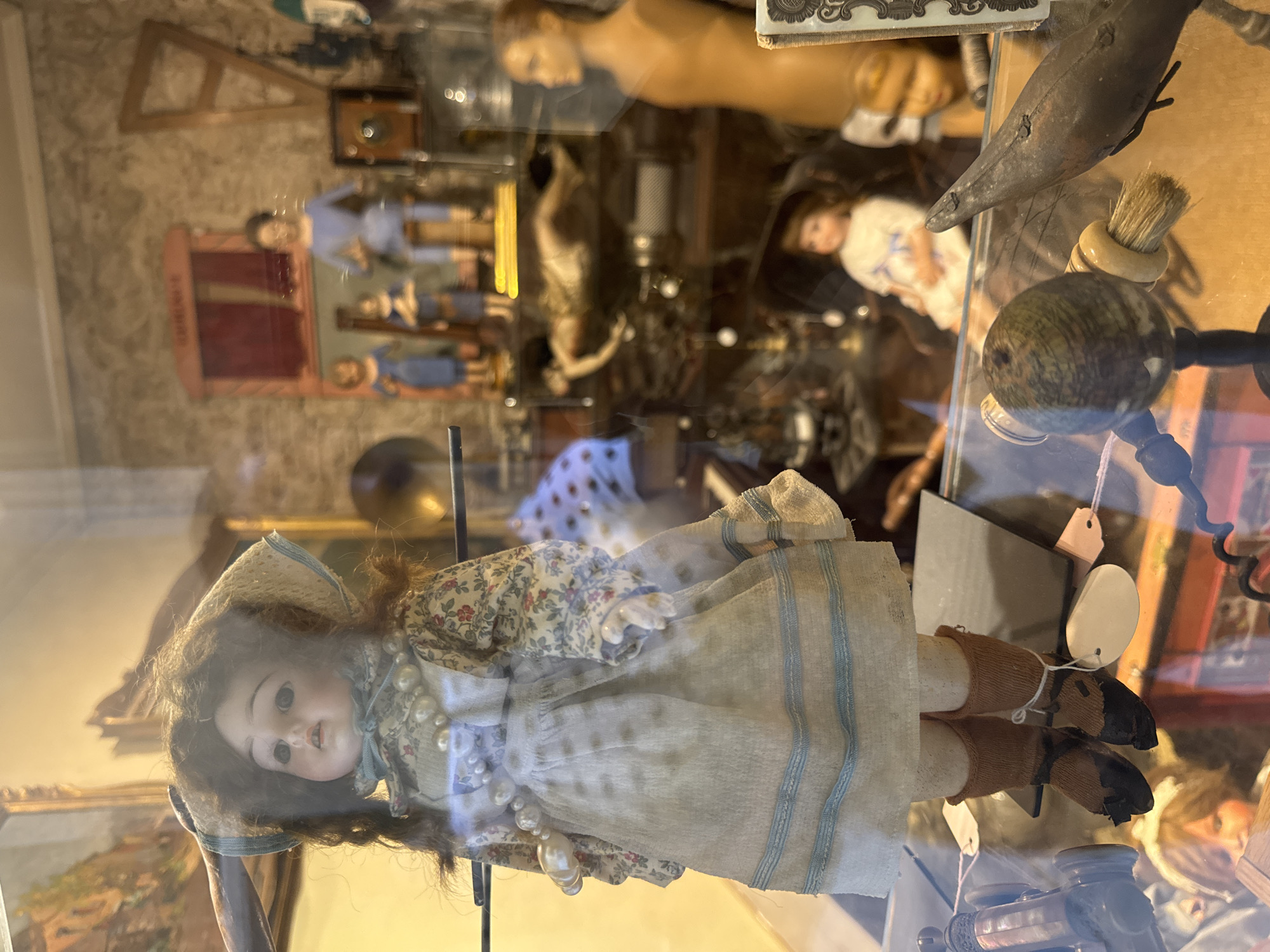 This vertical image depicts a detailed view into a glass display case, likely in an antique shop or museum in Barcelona, Spain. The primary subject is a vintage doll positioned prominently in the foreground. This doll has a porcelain-like face, dark curly hair, and large, dark eyes, wearing a straw hat, a floral-patterned blouse, a light-colored skirt with blue vertical stripes, and a long pearl necklace. Her feet are clad in brown socks or boots.
Behind the front doll, a variety of other objects and figures are visible, also behind glass, contributing to a sense of a curated collection. These include several other dolls or mannequins in various states of dress and poses; for instance, a bare-chested mannequin figure is seen, as well as a smaller doll dressed in blue. Further back, a wooden easel-like structure, a dark bird-shaped ornament, a small brush with a rounded handle, and a spherical wooden object are discernible. Reflections on the glass case are noticeable, slightly obscuring some background details. Small, light-colored tags, possibly price tags, are attached to some items. The scene captures a still life of nostalgic and antique objects in an indoor setting. No legible text is visible.