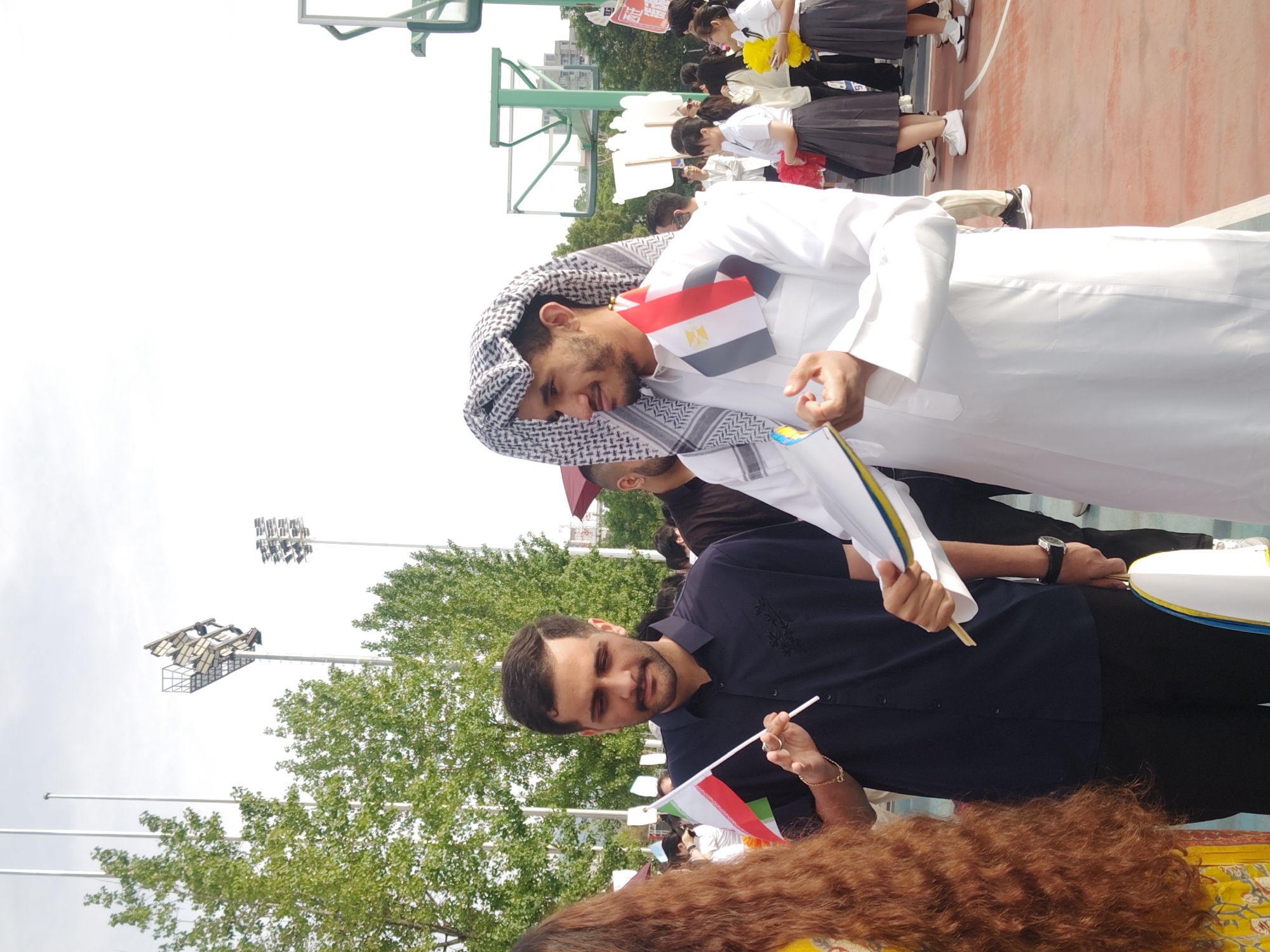 The image shows two men standing in an outdoor setting in Chongqing High-tech Zone, China. One man is wearing traditional Arab clothing, including a white thobe and a keffiyeh headscarf. He has an Egyptian flag draped over his shoulder. The other man is wearing a dark blue shirt and is holding a small Iranian flag. In the background, there are several people, some wearing school uniforms. There is a basketball hoop and trees visible, as well as some overhead lighting structures. The ground appears to be a sports court.