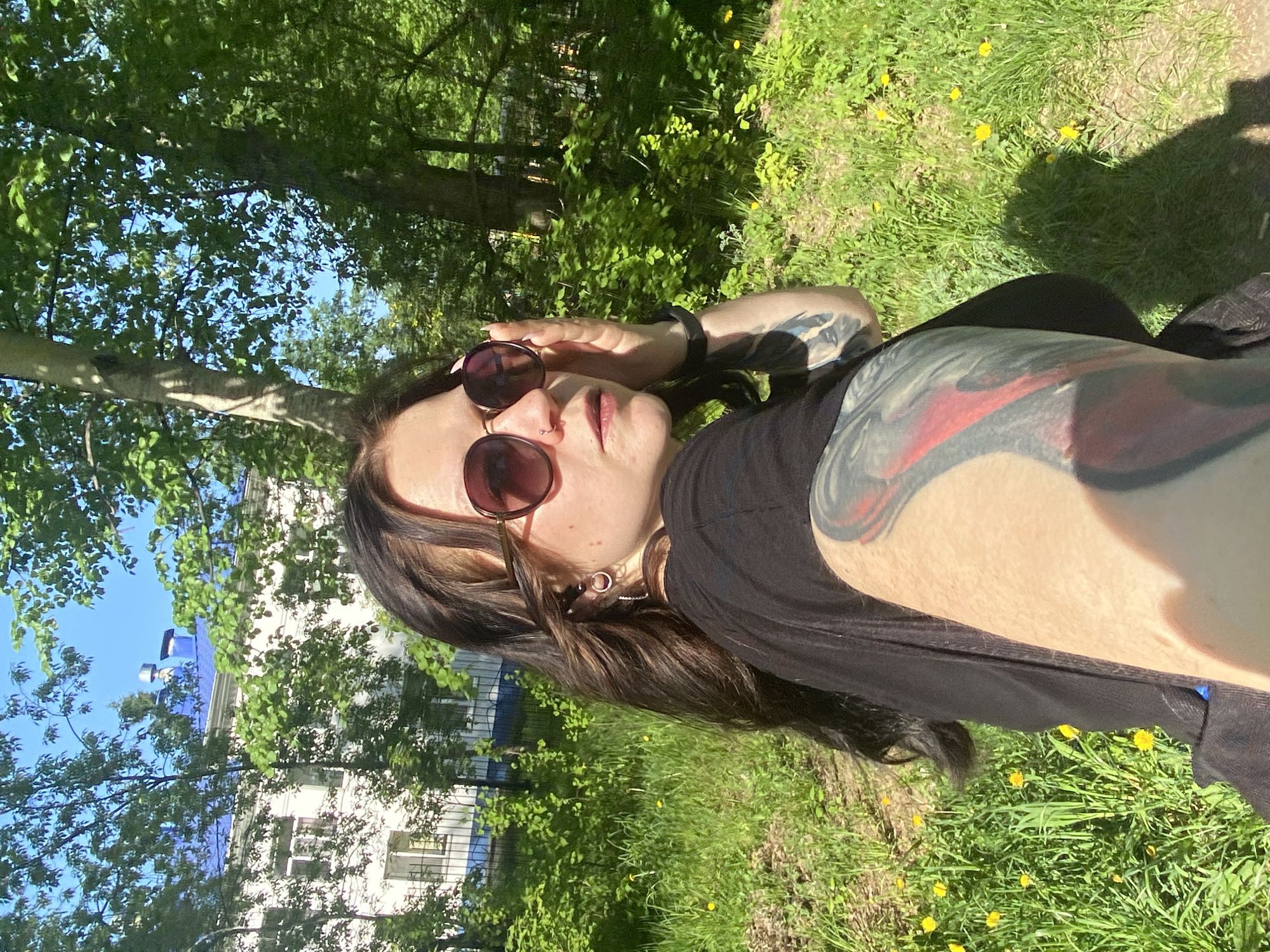 The image shows a person with long, dark brown hair and sunglasses, posing in what appears to be a grassy area with dandelions. The person has tattoos on their arm. In the background, there are trees and a building with a blue roof. The image appears to be taken outdoors, possibly in a park. Given the image's metadata, this is potentially within the city of Chaikovsky, Russia.