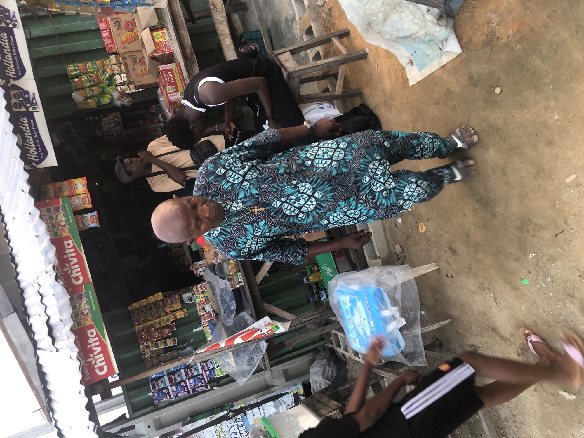 The image captures a street scene in Ado, Nigeria, depicting an outdoor market stall. In the foreground, an elderly man with a bald head, wearing a patterned blue and black tunic and pants, walks towards the viewer with a slight stoop. He is adorned with a gold cross necklace. To his left, a person wearing black shorts with white stripes and a black t-shirt is partially visible, walking away from the viewer. Their legs are blurred with motion, and they are wearing pink flip-flops. Behind the elderly man, a young man in a black sleeveless shirt with white trim is seated on a wooden stool, looking down at his phone. Another man in a light brown t-shirt and a dark cap stands at the market stall, appearing to be talking or eating. The market stall itself is laden with various products, including drinks, packaged snacks, and boxes. Prominent signage includes "Chivita" and "Hollandia." The ground is dirt and sand, with scattered debris. The overall atmosphere suggests a bustling, everyday scene in a Nigerian city.