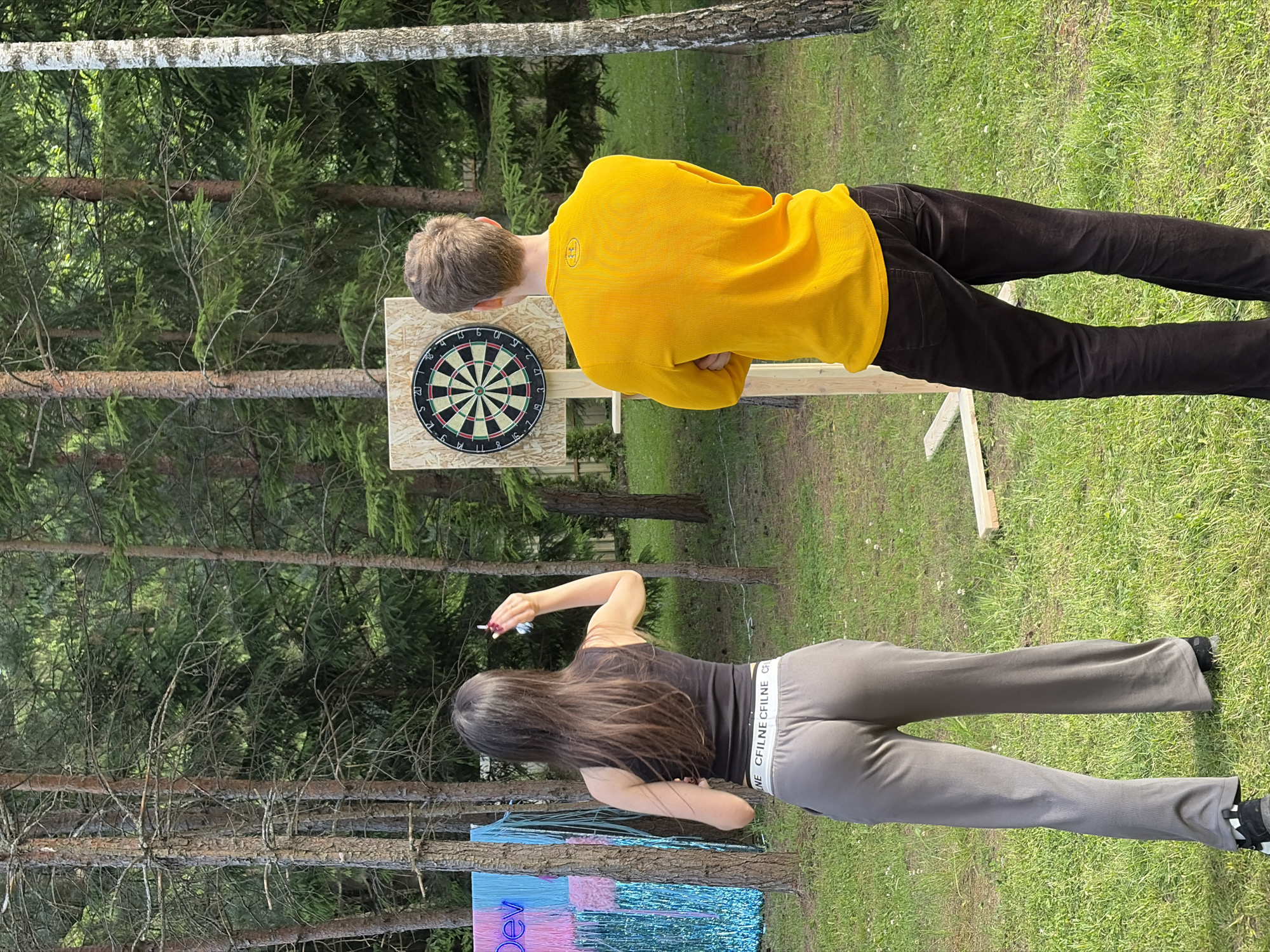 In the Ждановіцкі сельскі Савет, Belarus, a young woman with long brown hair, wearing a black tank top and grey sweatpants, stands facing away from the camera, holding a dart in her right hand, poised to throw. She is positioned to the left of a wooden dartboard mounted on a stand. To the right of the dartboard stands a young man, also facing away from the camera, wearing a yellow long-sleeved shirt and dark pants, with his arms crossed. The scene is set outdoors in a grassy area with numerous trees, suggesting a forest or park environment. The lighting indicates it is daytime, and the presence of a decorative blue and pink fringed backdrop with the word "Dev" visible on the left side of the image suggests a possible event or gathering.