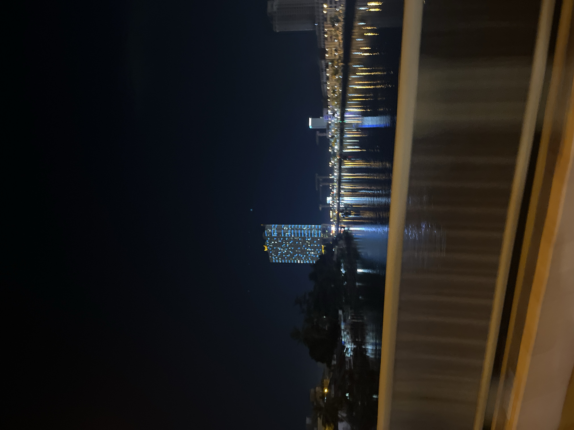 Nighttime view of a city waterfront in Nha Trang Ward, Vietnam, with the skyline reflected in the water. Buildings are visible in the background and along the water's edge. The picture is taken through a structure, possibly a window or railing, which obscures part of the scene on the right.