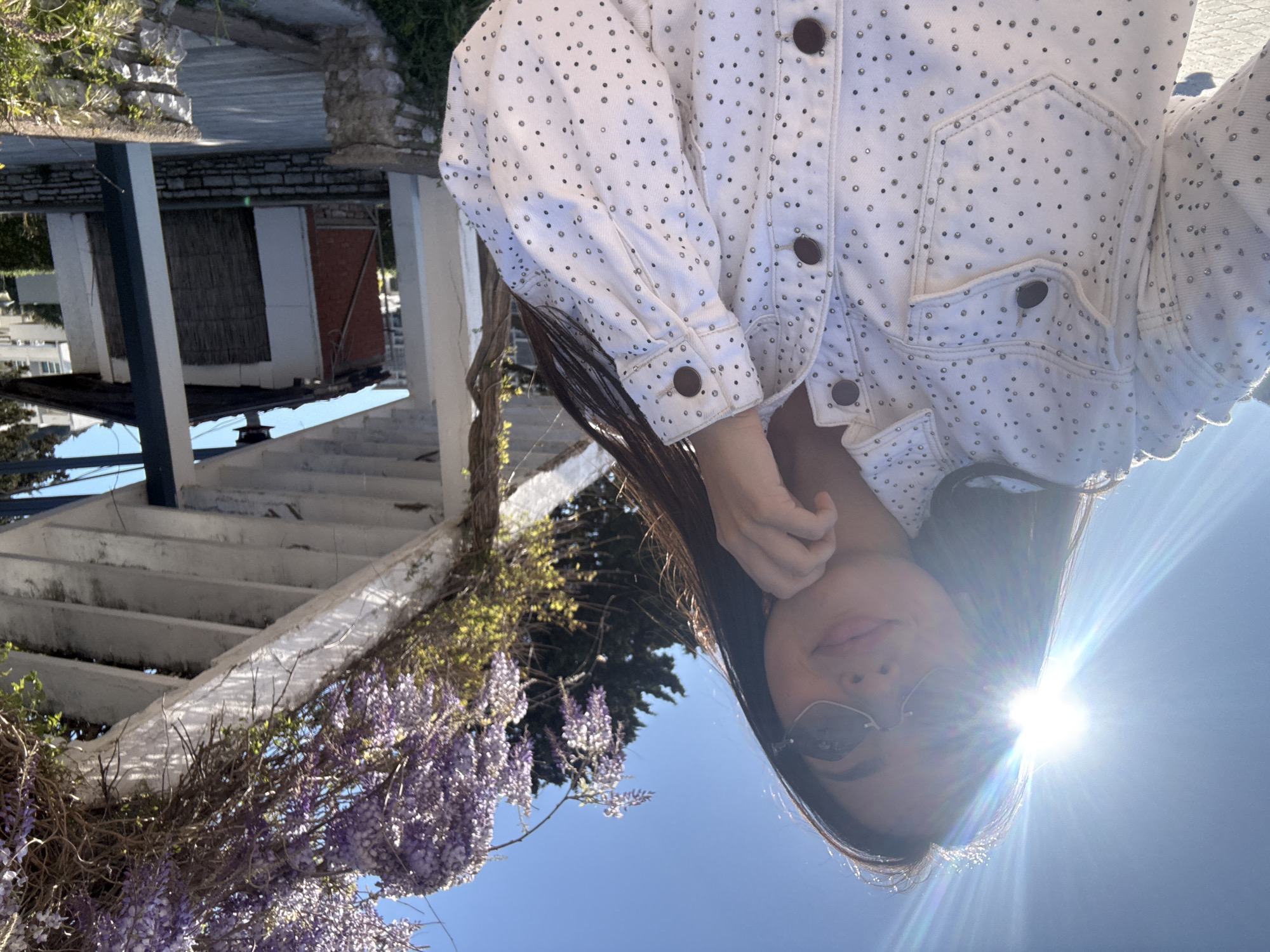 A woman wearing sunglasses and a white studded jacket is standing under a pergola structure covered in blooming wisteria. The background shows a clear blue sky and a building with a reflective surface, suggesting it may be near the sea. The bright sun is directly behind the woman's head, causing lens flare. The location is Unknown, Unknown.