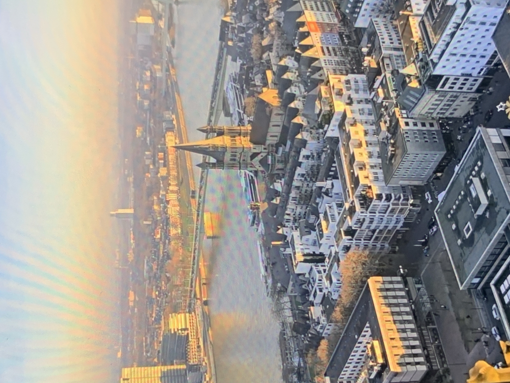 This is an aerial photograph capturing a cityscape bathed in the warm, golden light of sunset or sunrise. The image prominently features a large river flowing through the urban landscape, with bridges spanning its width. Dominating the foreground and midground are numerous buildings, a mix of architectural styles, some appearing modern with sleek facades, while others exhibit more traditional designs with pitched roofs. A significant structure resembling a cathedral with twin spires and a prominent clock tower stands out amongst the urban sprawl, its stone facade catching the warm light. The river is bordered by embankments and walkways, with several boats or ferries docked or traversing the water. In the background, the city extends further, with more buildings and a hazy sky, suggesting a broad metropolitan area. The overall impression is one of a bustling European city, likely on a river, with significant historical and modern architecture coexisting. The photograph does not depict Stavropol, Russia.