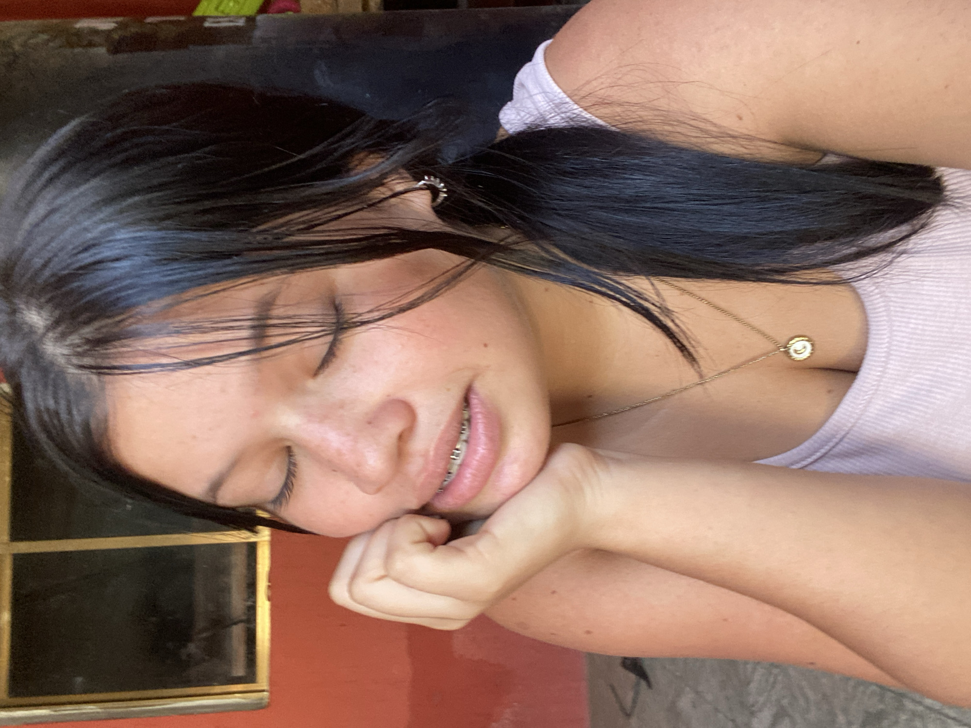 A woman with dark hair pulled back in a partial ponytail is resting her face on her hand. She has her eyes closed and is wearing braces. She is also wearing a light purple tank top and a gold necklace with a circular pendant. In the background, there is a window with a red frame. The location is likely indoors. Given the reference to Juan Griego, Venezuela, this is likely where the photo was taken.