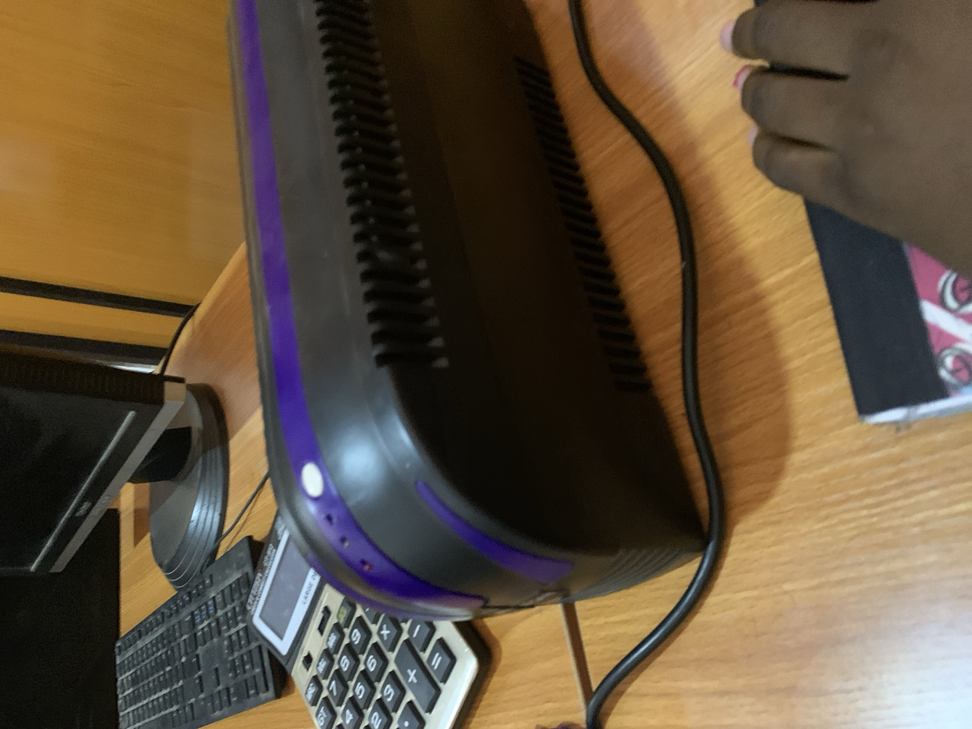 The image shows a close-up of a desk with various electronic devices. In the foreground, a black and purple device with a ribbed texture is visible. To its left is a silver calculator with a prominent display and keypad. Behind these items, a black keyboard is partially visible, along with the base of a computer monitor. The desk surface appears to be made of wood. A hand with pink fingernails is present in the lower right corner of the frame, resting on a black surface. The background is out of focus, showing what appears to be a wall with vertical lines. The overall setting suggests a home or office workspace.