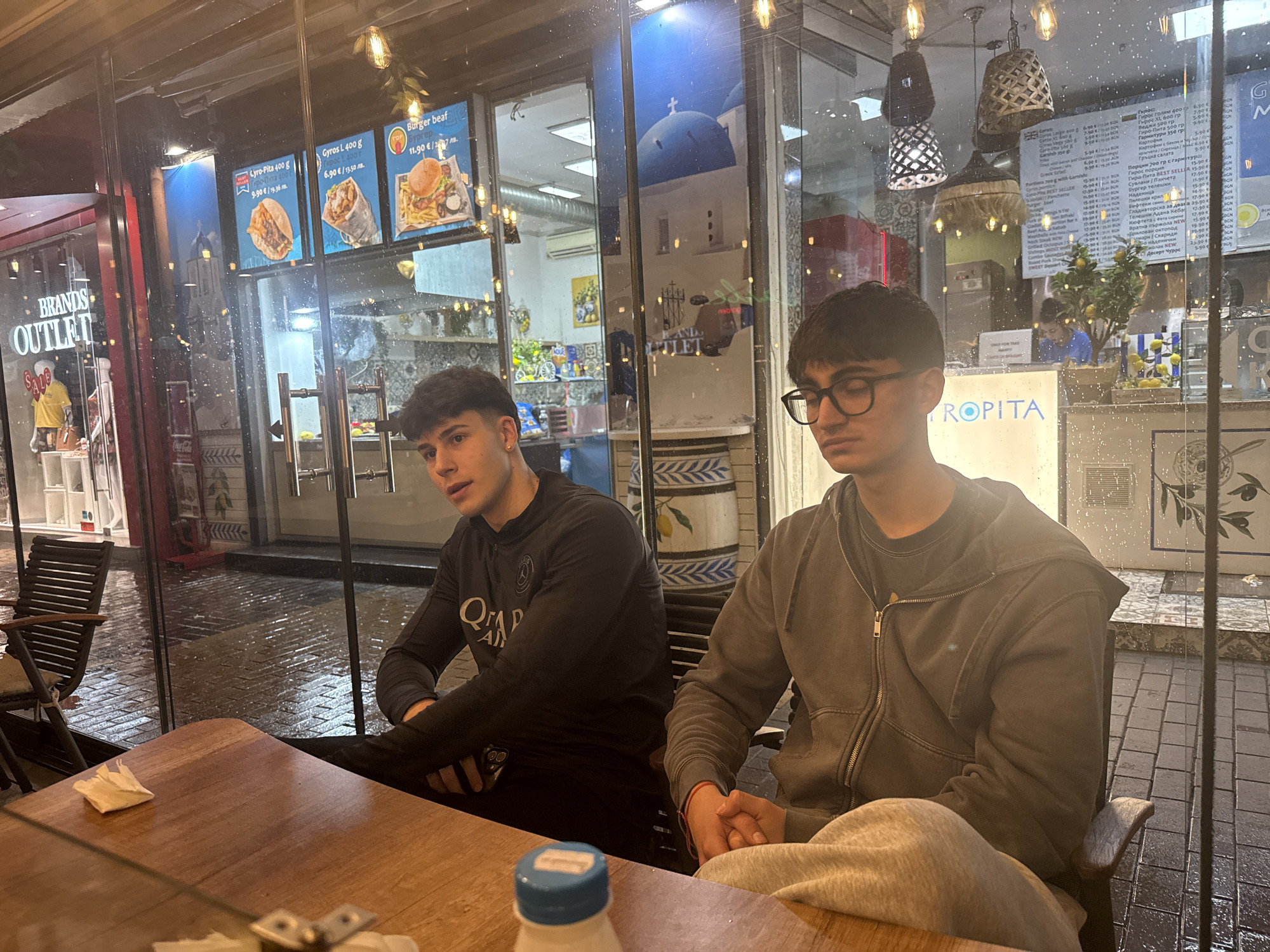 Two young men are seated at a wooden table inside a restaurant or cafe, looking out through a large glass window. The setting suggests evening or night, with artificial lights illuminating the interior and reflections on the wet glass from rain outside.
The man on the left has dark hair and wears a black long-sleeved shirt with a partially visible "QATAR AIRWAYS" logo. He looks off to his left with a pensive expression. The man on the right, also with dark hair, wears glasses and a grey zip-up hoodie over a dark t-shirt. His eyes are closed or cast downwards, giving him a relaxed or introspective appearance. On the table between them, a white napkin and a white bottle with a blue cap are visible.
Through the window, several details are apparent. The glass is covered in raindrops, and the pavement outside appears wet, confirming recent rain. Reflected in the glass are images suggestive of Greek architecture, including a distinctive blue dome and white buildings. Menu boards display food items such as "Gyros-Pita 400 g," "Gyros L 400 g," and "Burger beef," with prices listed in both Euros (€) and Bulgarian Levs (лв.), consistent with the Sofia, Bulgaria location. Other menu items in Cyrillic script are also visible, including "Гирос", "Бургер телешки", and "Десерт Чуррос". A shop sign for "BRANDS OUTLET" is reflected outside, and a partial sign for "TROPITA" is visible inside. Decorative elements like a small lemon tree and traditional-looking tiled patterns further enrich the scene. The overall ambiance is casual and somewhat intimate, with the two individuals seemingly pausing from their activities indoors while observing the rainy street outside.