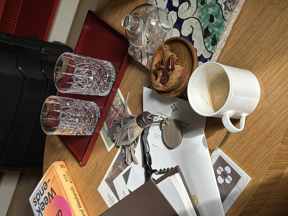 The image displays a collection of items arranged on a light wooden table, indicative of a hotel room setting in Paris, France, specifically associated with the "Experimental Hotel" brand. No individuals are visible.
In the foreground, a white ceramic mug containing a light brown beverage stands next to a small wooden plate holding a partially consumed circular dessert topped with chocolate and pecans. Adjacent to the dessert is a clear glass cloche. A decorative tile with an abstract blue and green pattern lies partially beneath these objects.
Centrally positioned on the table are various papers, including a document labeled "HORAIRES DU RESTAURANT" and a small card reading "HOTEL EXPERIMENTAL." A black corkscrew and a beige leather keyring with a tassel and the number "50" are interspersed among these papers, along with a dark brown notebook.
Further back, a red lacquered rectangular tray holds two clear, patterned glass tumblers. To the left, an open book with an orange cover, titled "Weekends" by "DISIN MCKENNA," is visible. The background reveals a dark, ribbed object consistent with luggage or furniture. The scene implies recent guest occupancy and use of the presented amenities.