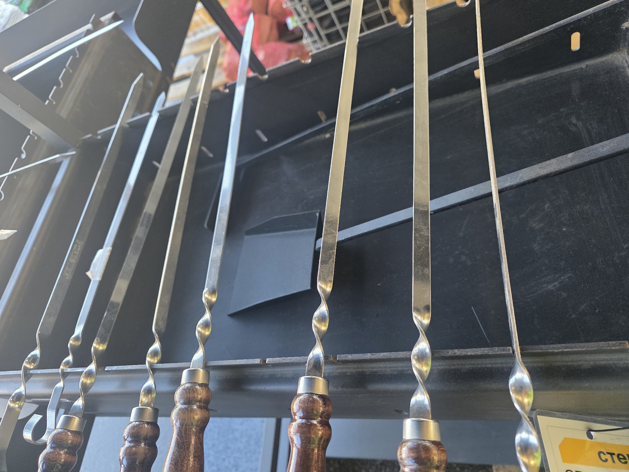 A close-up, low-angle shot shows several metal skewers with twisted shafts and wooden handles, resting on a black grill. The skewers are arranged in a row, with their pointed ends facing upwards and slightly out of focus. The twisted design of the metal shaft is visible on each skewer, as is the dark, polished wood of the handles. A black grill, with visible metal grates and supports, forms the background. The overall scene is outdoors, with natural light illuminating the skewers and grill. There are no people or animals visible in the frame.