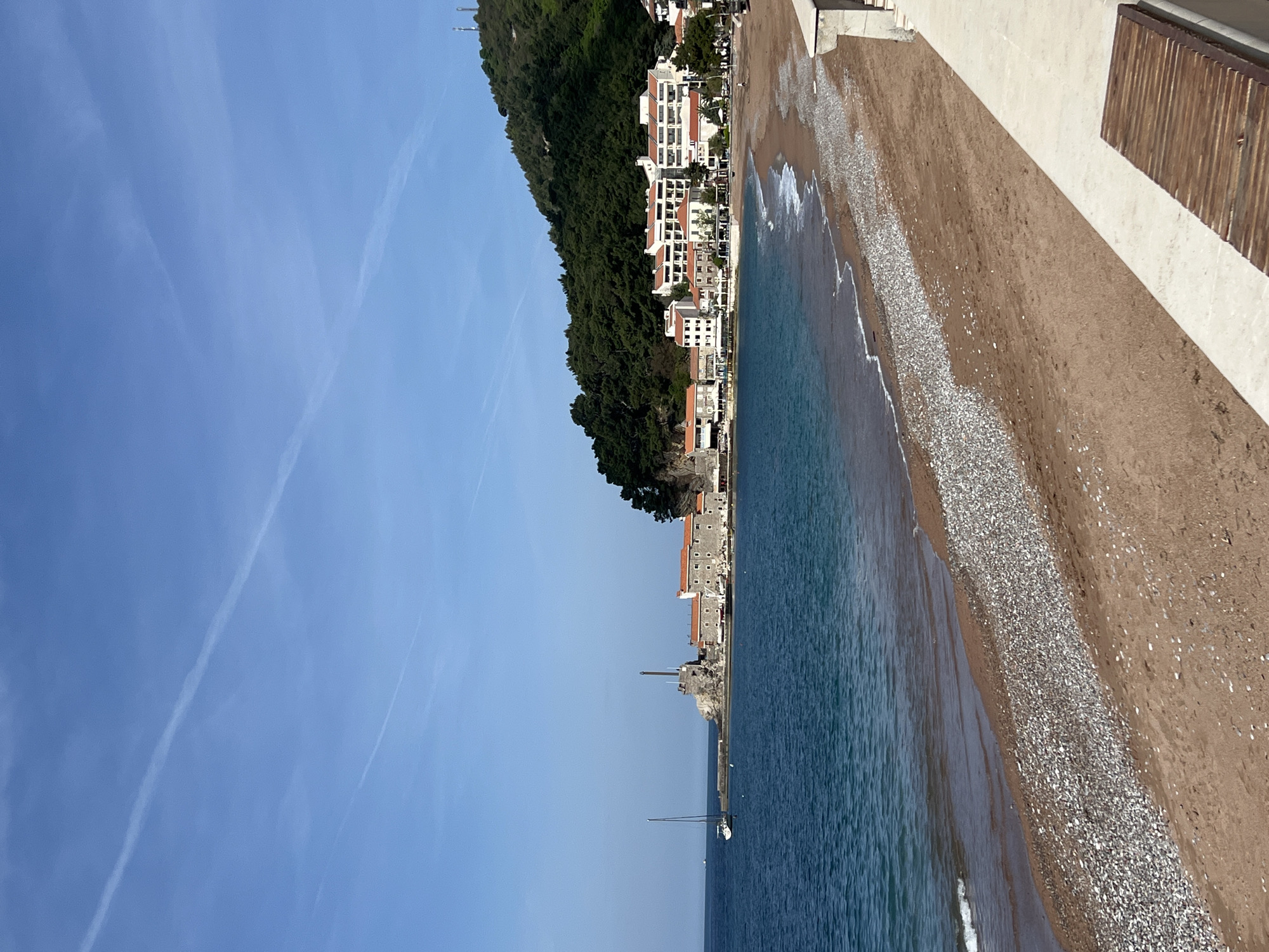 The image depicts a coastal scene in Petrovac centar, Montenegro. The view is oriented along the shoreline, showing a beach with a mix of sand and pebbles meeting the clear blue water of the Adriatic Sea. Waves gently lap the shore. To the left of the beach are several multi-story buildings with white facades and red-tiled roofs nestled against a densely wooded hillside. The sky above is bright blue with faint contrails suggesting recent air traffic. A single sailboat is visible in the water. The scene suggests a tranquil seaside resort environment.