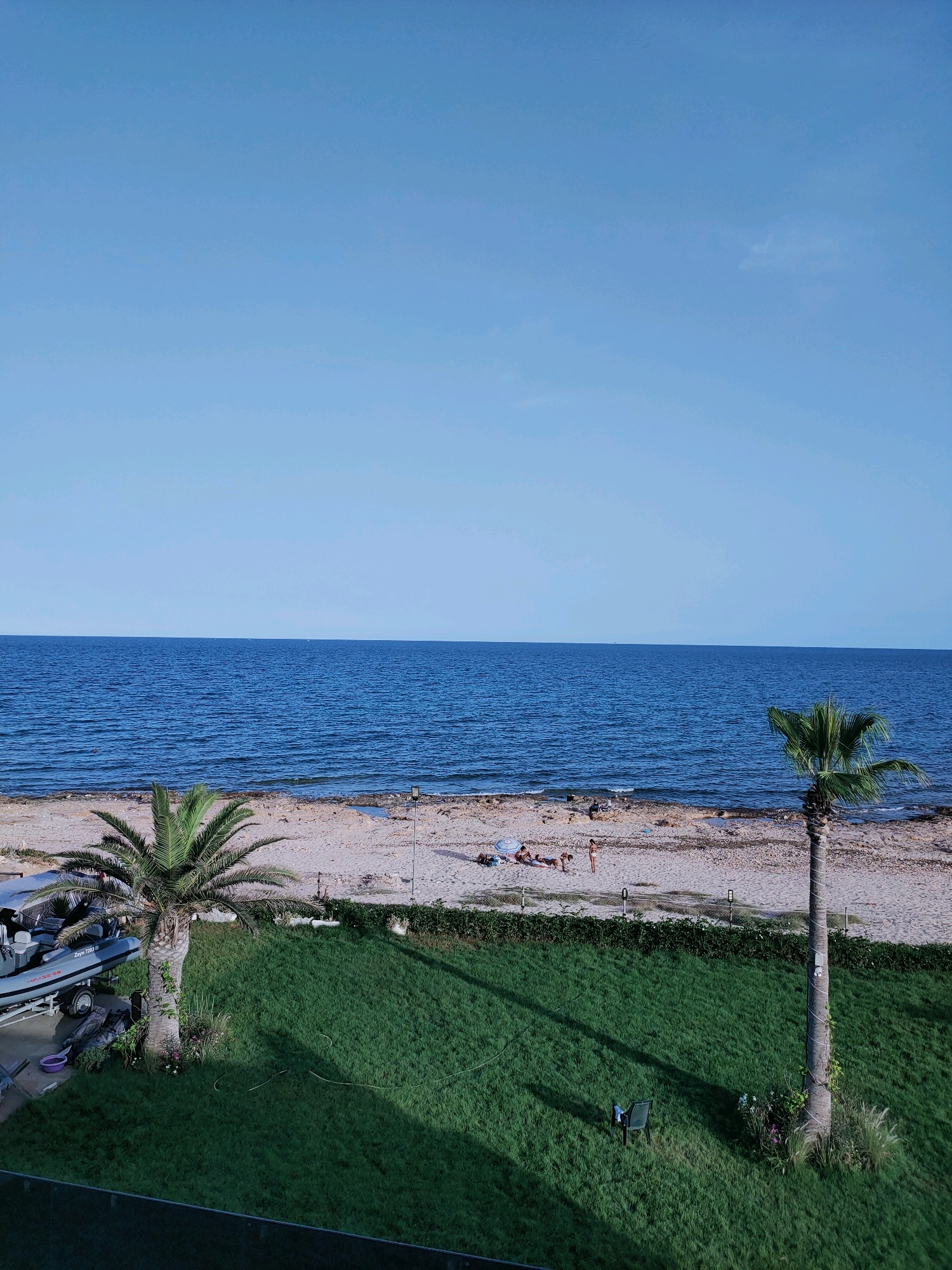 An aerial shot captures a serene beach scene in Maâmoura, Tunisia. The clear, bright blue sky dominates the upper portion of the frame. Below, the deep blue sea stretches horizontally, meeting the shoreline in a gentle wave pattern. The beach itself is a mix of sand and rocks, with several people visible sunbathing and relaxing. Two palm trees stand on the beach, one on the left and another on the right, adding to the tropical ambiance. In the foreground, a well-maintained green lawn is visible, with a small chair casting a shadow. To the left of the lawn, a boat on a trailer is parked, suggesting a recreational area. The overall scene is peaceful and indicative of a sunny day by the Mediterranean Sea.