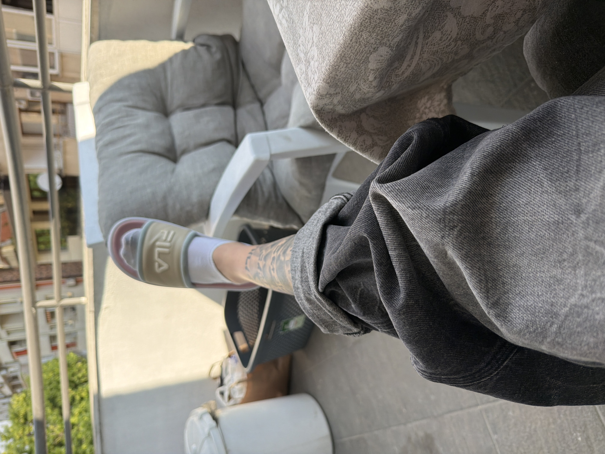 The image captures a close-up view of a person's legs and feet, adorned with a Fila slide sandal over a white sock. The person is wearing dark, distressed denim jeans, which are casually draped. The scene appears to be on a balcony or outdoor seating area, with a grey upholstered chair and a table covered with a patterned cloth visible in the background. A white cylindrical object, possibly a trash bin, and a dark grey laundry basket are also present. In the distance, buildings and trees suggest an urban setting, likely within the city of Plovdiv, Bulgaria, given the architectural style of the buildings. The lighting indicates it is daytime, with sunlight casting shadows.