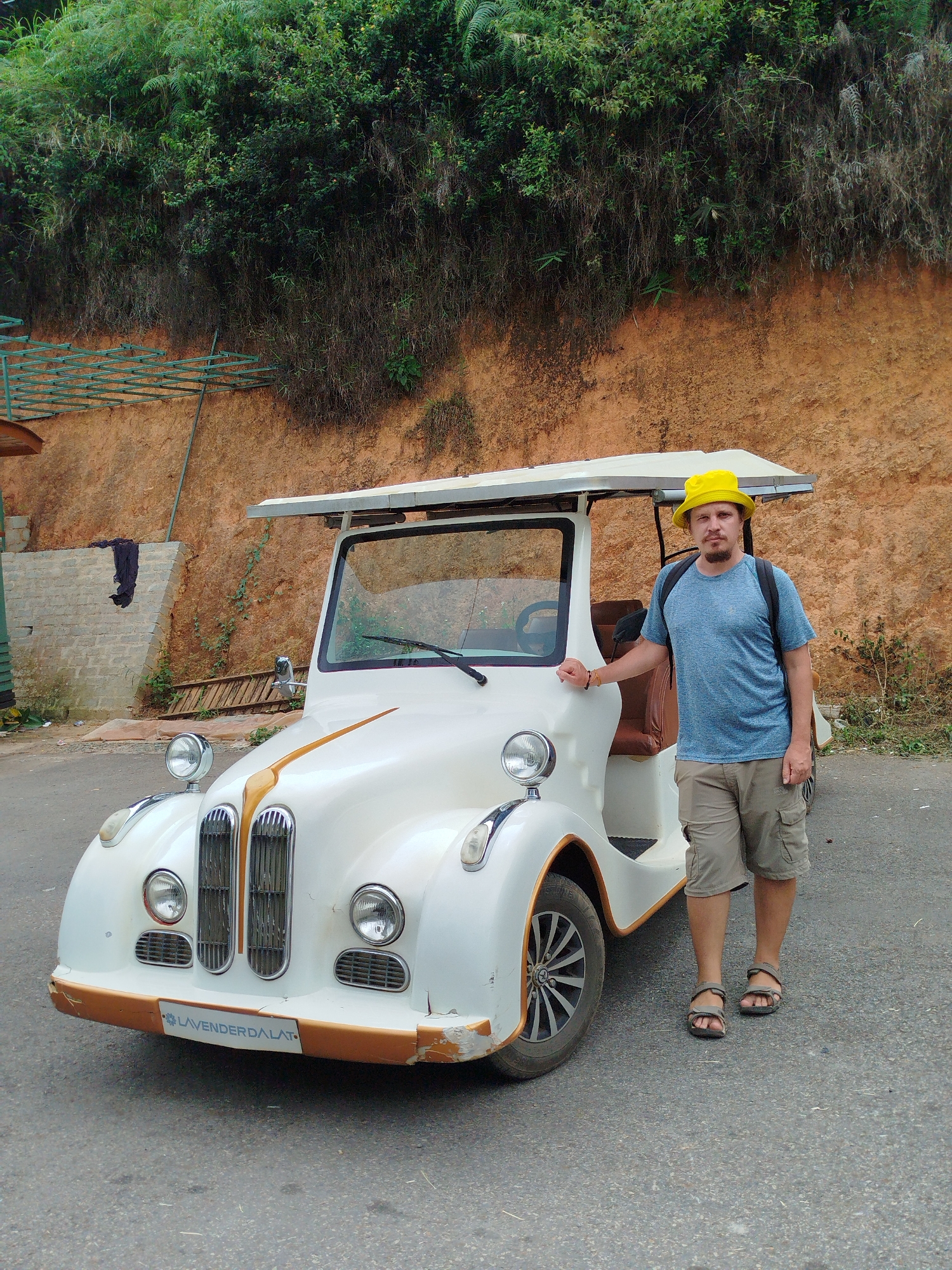 In Da Lat, Vietnam, a man wearing a yellow bucket hat and a blue t-shirt stands next to a vintage white golf cart. The golf cart has a tan interior and a light-colored canopy. The man's left hand rests on the side of the golf cart, and he is wearing tan cargo shorts and sandals. The background consists of a steep, earthy embankment covered in lush green foliage and vegetation. A ladder leans against the embankment to the left of the golf cart, and a dark piece of fabric is hanging from a structure on the left. The ground is paved, suggesting a road or parking area. The license plate on the golf cart reads "LAVENDERDALAT".