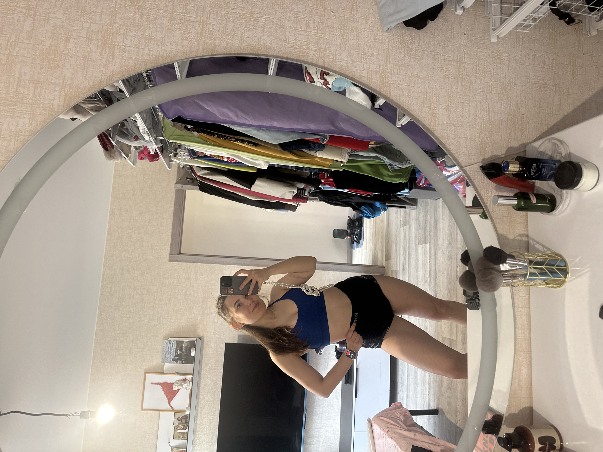 A young woman is taking a mirror selfie in what appears to be a bedroom or dressing area. The image is originally rotated 90 degrees counter-clockwise, showing the woman standing upright, reflected in a large, irregularly shaped mirror with a wide, frosted-looking frame.
The woman has long brown hair and is wearing a blue sports bra and black shorts with "BBO" or "BBG" visible on the waistband. She accessorizes with a long pearl necklace draped around her neck and a dark smartwatch on her left wrist. She holds a dark-cased smartphone in her right hand, angled to take the photo, while her left hand rests on her hip. She has a slight smile and looks directly at the camera in her phone.
The reflection in the mirror reveals a room with light-textured wallpaper and light wood-look flooring. Visible elements include a large flat-screen television on a low white cabinet, a shelf with several framed pictures (one prominent red abstract piece), and a single exposed light bulb hanging by a wire. Behind the woman, an open closet or clothing rack is filled with various garments in different colors (purple, green, yellow, black, white, red, denim). Further in the background, a piece of black exercise equipment is partially visible.
Outside the mirror's reflection, to the right, a white surface holds several items. These include a dark green spray bottle, a red tube, a blue container labeled "BLUE TALISMAN" (partially visible text like "DR. HO..." is also present), and a white jar. Adjacent to these, a gold geometric-patterned holder contains several fluffy makeup brushes. The overall setting suggests a personal and lived-in space.
