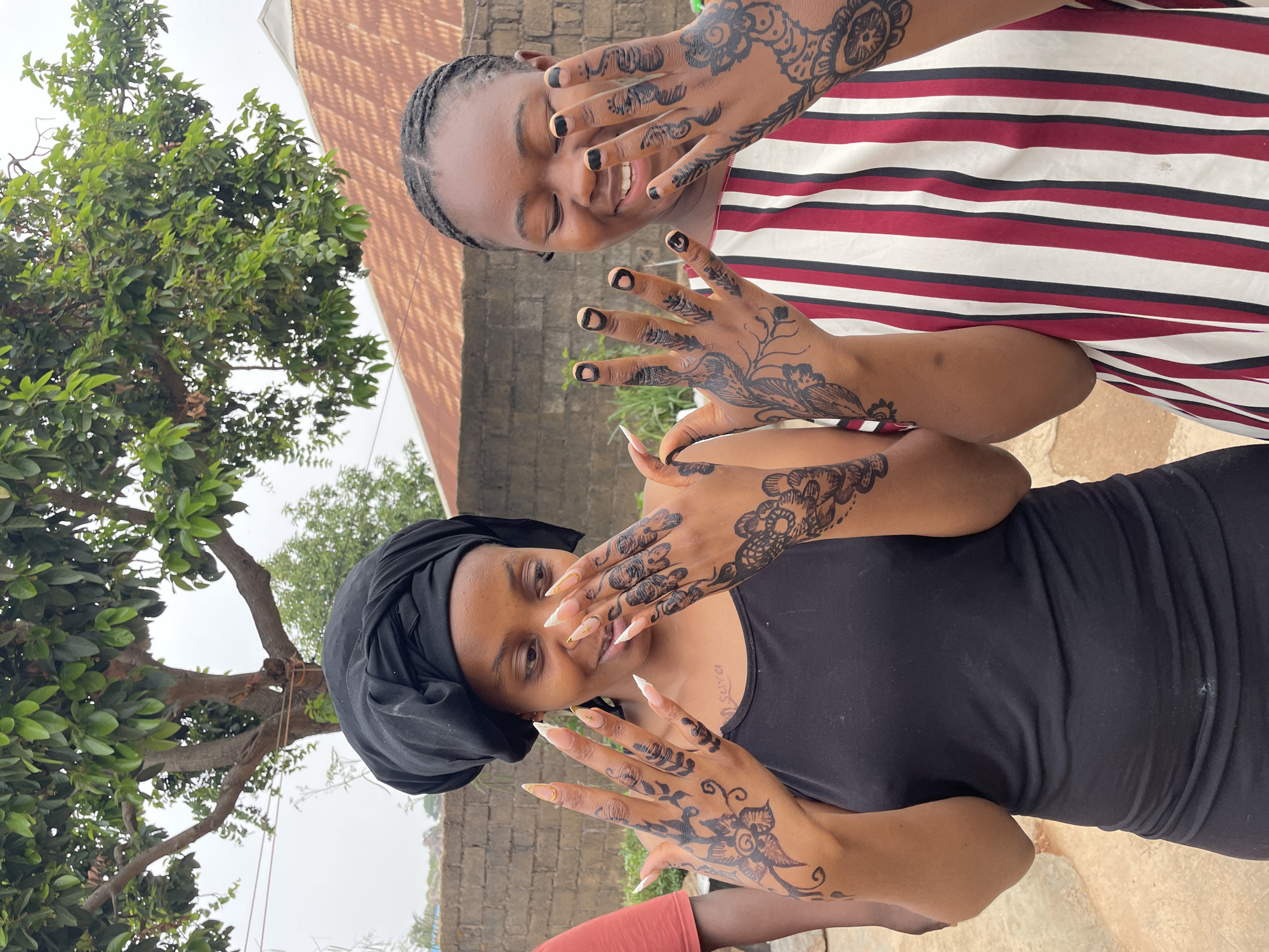 Two women are posing for a photograph, showcasing henna designs on their hands. The woman in the foreground wears a black head covering and a black top, displaying long, pointed artificial nails. The woman behind her has braided hair and wears a striped red, white, and black top. The background includes a tree with green foliage, a low brick or concrete wall, and a building with a light-colored roof. The image was taken in Jos, Nigeria.