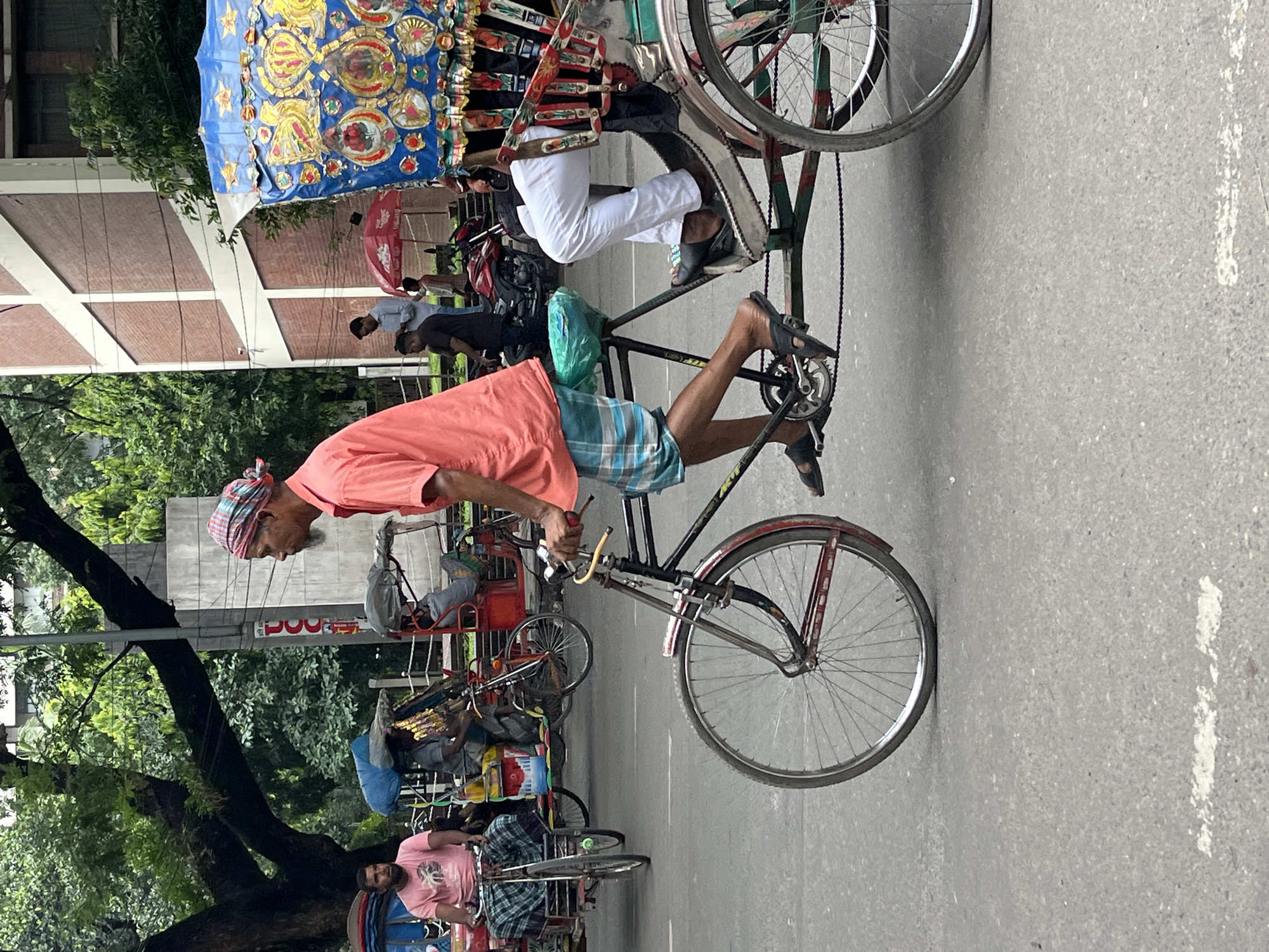 A rickshaw puller wearing a pink shirt and checkered lungi is pedaling a rickshaw on a street in Dhaka, Bangladesh. The rickshaw is decorated with blue and gold fabric. In the background, another rickshaw with passengers can be seen, along with buildings and trees. The street is paved and appears to be busy with traffic.