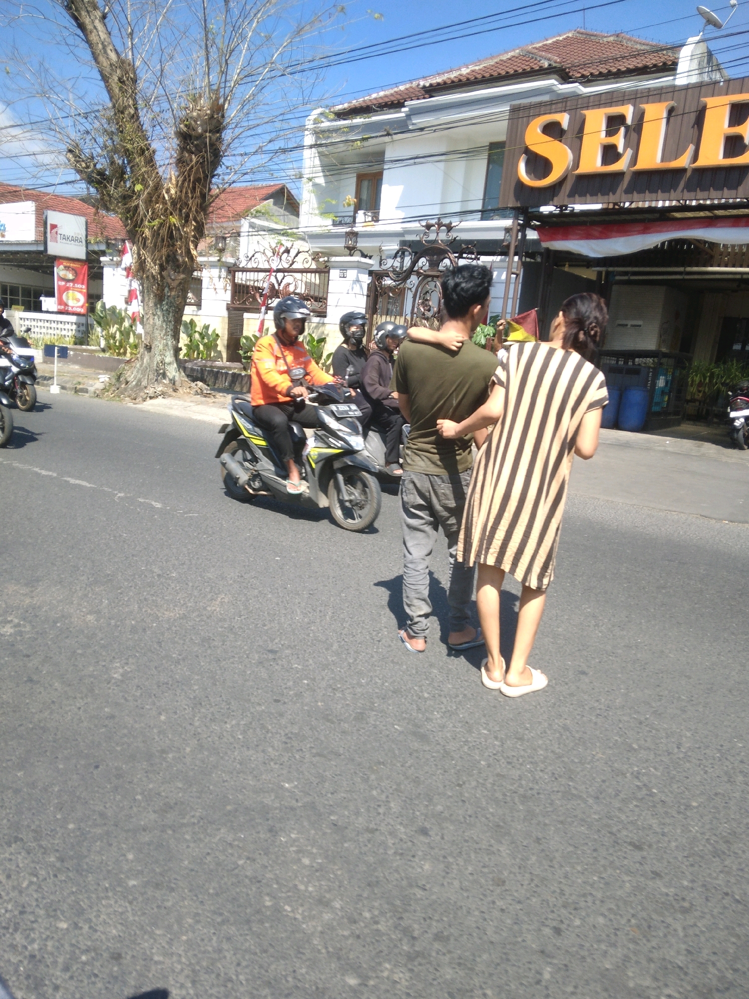 In Bancarkembar, Indonesia, on a bright, sunny day, two individuals stand on a paved road in front of a building with a large sign that reads "SELECT". The person on the left is a male wearing a dark olive-green t-shirt and gray pants, with his left arm around the shoulders of the person on the right. The person on the right is a female wearing a vertically striped dress in alternating shades of tan and dark brown. She is facing away from the camera. A motorcycle is visible to the left of the couple, and another motorcycle with a rider wearing an orange jacket and black helmet is in motion on the road to the left of the frame. In the background, there is a white building with a red-tiled roof and an ornate gate. Trees with bare branches are also present. The road is asphalt, and there are white painted lines indicating a lane.