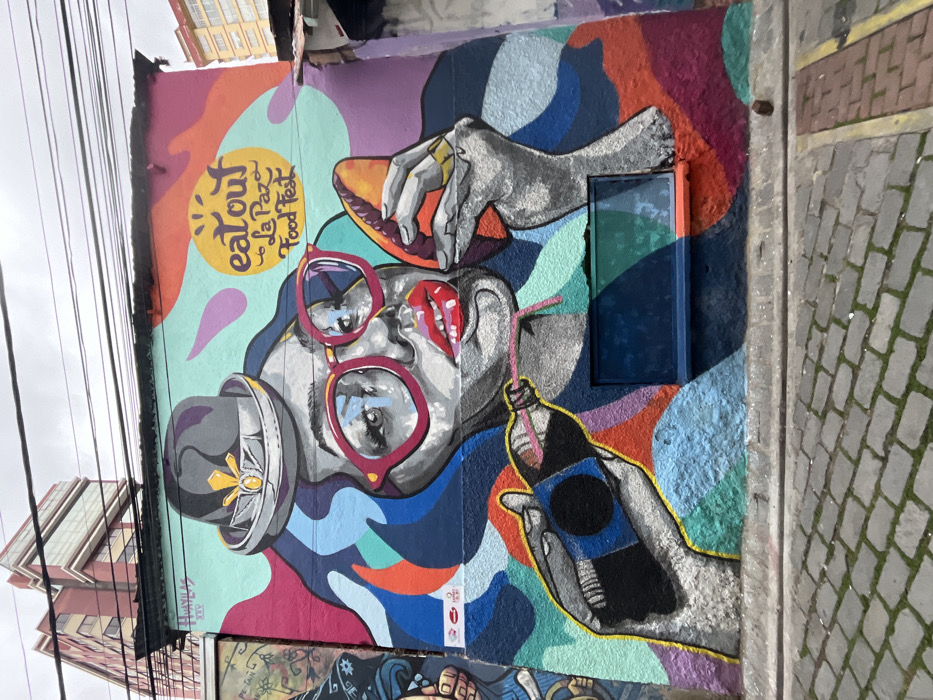 A vibrant mural features a person wearing large red sunglasses and a hat, seemingly enjoying food and drink. A hand holds a sandwich or empanada above their head, while another hand presents a bottled beverage with a straw. The mural is colorful with a mix of abstract shapes and patterns in shades of blue, orange, purple, and green. The text "eat out La Paz Food Fest" is also visible. A gray brick wall and building are to the right and left of the mural, respectively.