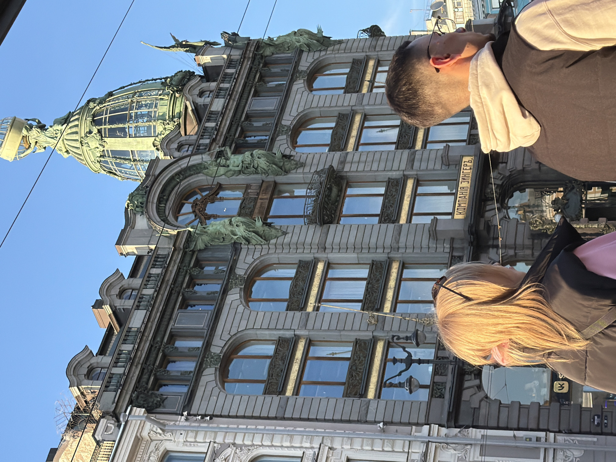 The image depicts the Singer House in Saint Petersburg, Russia, as seen from below. The building's ornate facade, including the dome, is visible. Two individuals are partially in the foreground, their faces obscured. The sky is visible in the background.
