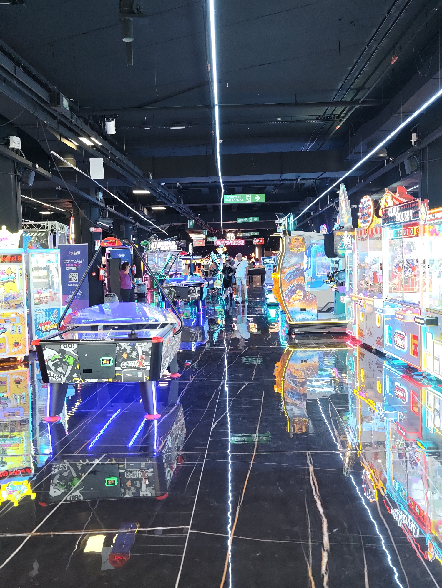 This image depicts the interior of a modern arcade and entertainment center in Nola, Italy. The space features a high ceiling with industrial lighting fixtures and ventilation systems. The floor is polished black marble with white veining, highly reflective and mirroring the bright neon lights and arcade machines.
The foreground is dominated by an air hockey table with blue LED lighting accents and colorful graphics. Behind it, a row of various arcade machines is visible, including claw machines with prizes inside and video game cabinets with vibrant digital displays. One machine prominently displays the text "GAME ORBIT" and instructions in Italian.
In the mid-ground, several people are present. A woman wearing a purple shirt is standing near a fire extinguisher, looking at one of the arcade machines. Further back, a man in a white t-shirt and shorts is walking with a child.
The environment is characterized by a playful and energetic atmosphere, suggested by the flashing lights, colorful machines, and the presence of patrons. The overall impression is a well-lit, clean, and technologically advanced entertainment venue.