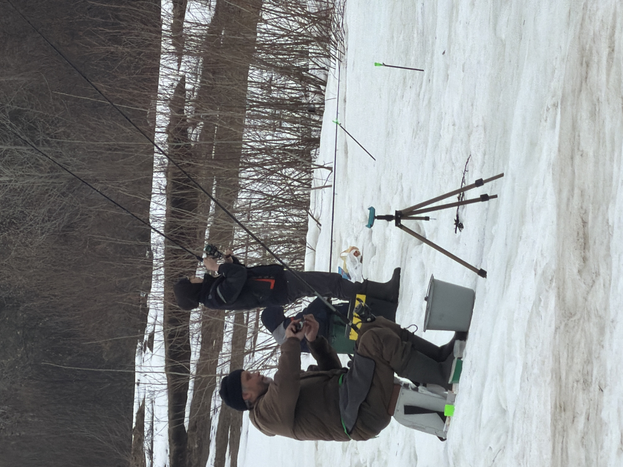 Two men are ice fishing on a snowy riverbank. The man in the foreground is wearing a brown jacket, dark pants, and a black hat and is reeling in a fishing line. The man in the background is wearing a black jacket and dark pants, also reeling in a fishing line. Both men are wearing rubber boots. A tripod with a blue object attached is near the snowbank. A grey bucket and a white container are located by the snowbank. Several thin sticks with green ends are stuck in the snow. The location is Chaikovsky, Russia, as the bank of the Kama River is a popular fishing location. Trees are visible on the riverbank.