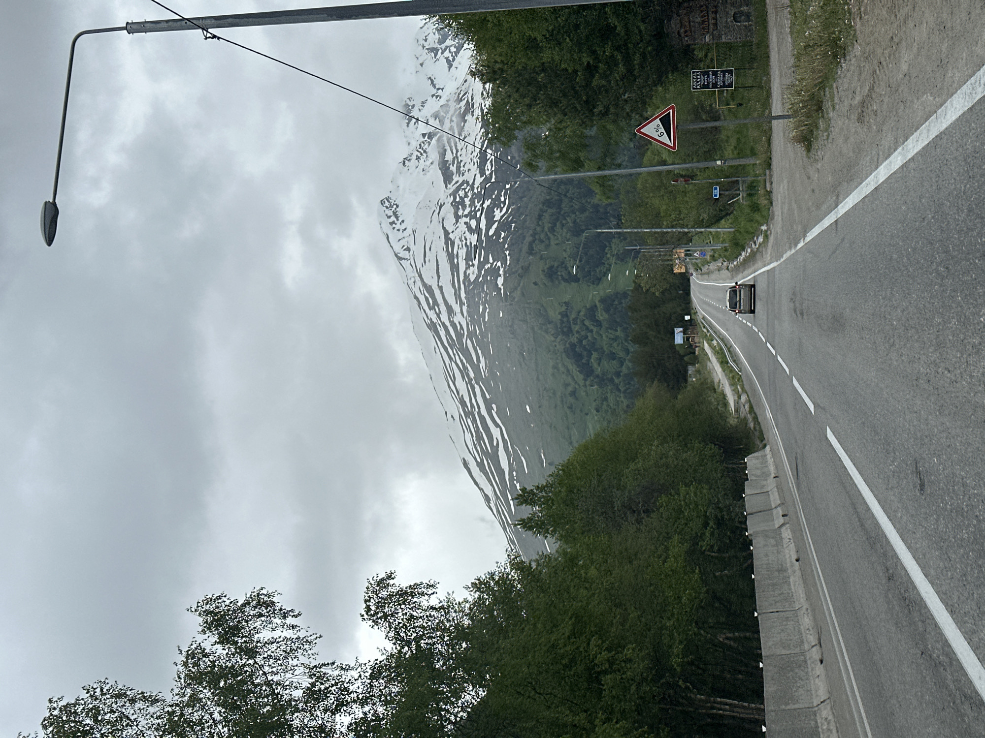 The image depicts a grey, overcast day in the mountainous region near Baidaevo, Russia. A paved road, marked with white lines, curves towards a snow-capped mountain in the distance. Trees line both sides of the road, with a concrete barrier on the left. A single car is visible driving on the road. A street lamp stands on the right side of the road, near a triangular sign indicating a 6% incline. Several other road signs are also present. The mountain in the background is partially covered in snow and has green slopes.