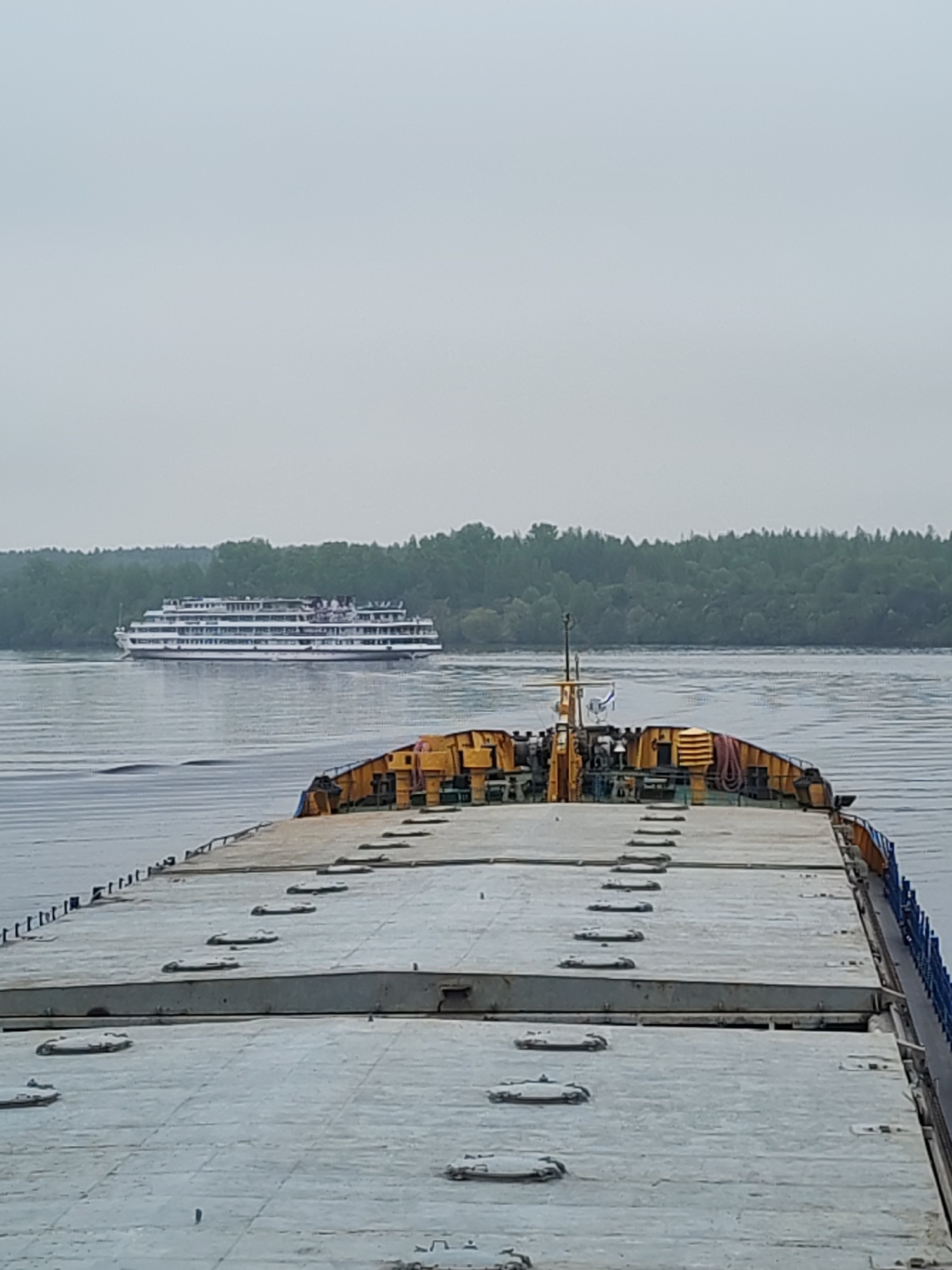 The image shows a water scene in Kalyazin, Russia. In the foreground is the flat deck of a barge, painted gray and with numerous access hatches. Behind the barge, on the water, is a white passenger ship with multiple decks and windows. On the horizon, there's a treeline with lush green trees, and the sky is overcast and gray.