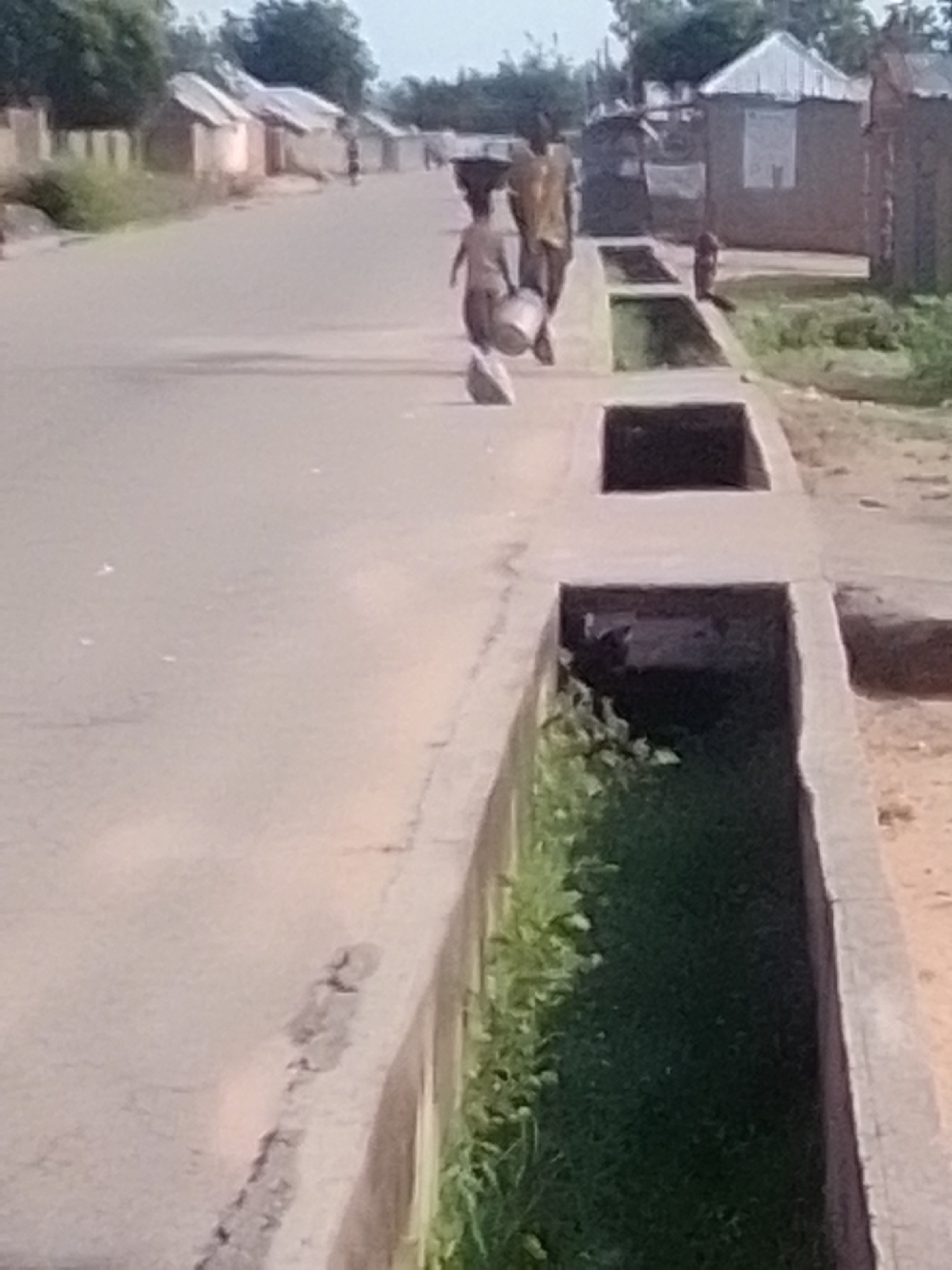 A young child with a bucket on their head walks down a paved road in Talasse, Nigeria. The child is accompanied by an adult, who is also carrying an object on their head. The road is lined with buildings on both sides, and there is a concrete drainage ditch running alongside the road. The environment appears to be dry and dusty, with sparse vegetation. The sky is overcast.