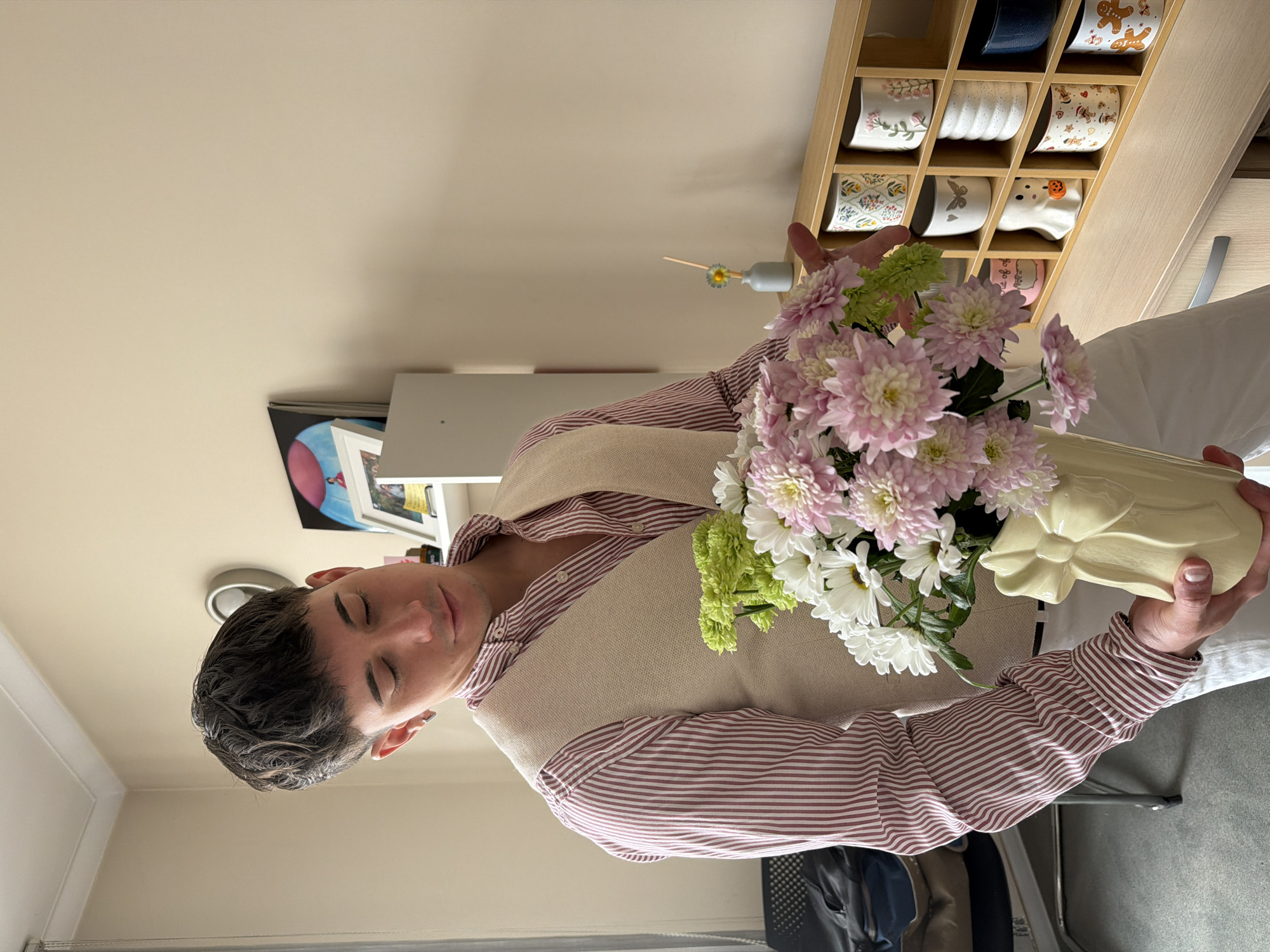 A man stands indoors, holding a vase of flowers. He wears a horizontally striped red and white shirt under a tan vest and white trousers. The vase contains pink, white, and green flowers. Behind him is a beige wall with a framed picture on it, a light grey storage unit, and a wooden shelf containing multiple mugs or cups. He is indoors. The location is not discernable to be Sofia, Bulgaria.