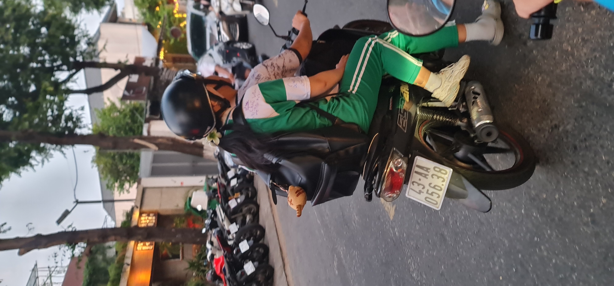 The image captures a street scene in Đà Nẵng, Vietnam, featuring two individuals on a black motorbike, likely during late afternoon or evening as indicated by the ambient light and illuminated building lights.
The prominent subject is the passenger of the motorbike, who is wearing a black helmet, a green and white tracksuit, and white sneakers. This person has long, dark hair with a white flower tucked into it, and carries a black backpack from which a small, conical, light brown charm or toy hangs. The driver of the motorbike is partially visible, wearing a white and black patterned shirt.
The motorbike itself is a black scooter-style model with a clear license plate displaying "43-AA 056.38" (43 is the provincial code for Đà Nẵng). Partial branding, possibly "HONDA" and "ES", is visible on its side. The exhaust pipe and tail light are also distinguishable.
The setting is an urban street with an asphalt surface. The background includes several trees with green foliage, buildings with glowing lights, and a street light pole. A line of other parked motorbikes is visible further down the street, consistent with typical transportation in Vietnam. The sky appears overcast or transitioning to night. Some indistinct writing is present on the back of the passenger's shirt.