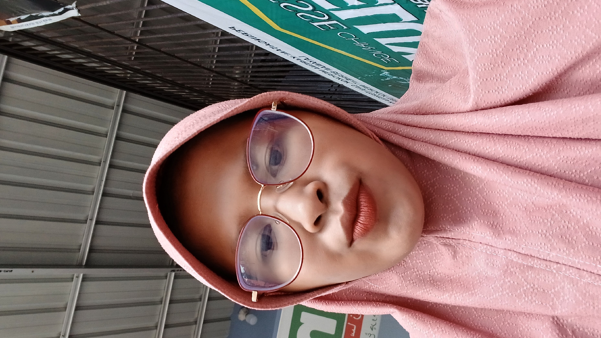 A woman wearing a pink hijab and glasses is captured in a close-up shot. She is positioned against a background that includes a corrugated metal structure and a green and white banner with Indonesian writing. The location is presumed to be in Lhokseumawe, Indonesia.