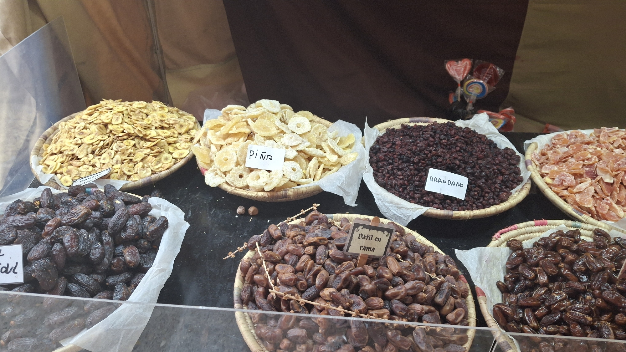 The image displays a market stall in La Fresneda / la Freixneda, Spain, featuring a variety of dried fruits. In the foreground, two woven baskets hold dates. The basket on the left contains darker, wrinkled dates, with a white tag visible. The basket to its right holds lighter brown, oval-shaped dates and has a wooden sign with the text "Dátil en rama" attached. To the left of these date baskets is a clear plastic container filled with dark, oval dates, with a partial white tag visible. Behind the dates, there are several more baskets of dried fruit. The basket furthest left contains a large pile of dried banana chips, with a white tag that is partially obscured. Next to it, a basket is filled with dried pineapple rings, identified by a white tag with "Piña" written on it. To the right of the pineapple, a basket is overflowing with dark red dried berries, labeled "ARANDANO" on a white tag. Further to the right, a basket contains lighter orange-colored dried fruit pieces. In the upper background, a brown cloth drapes behind the stall, and a few colorful lollipops are visible. The overall impression is that of a produce market or specialty food vendor.