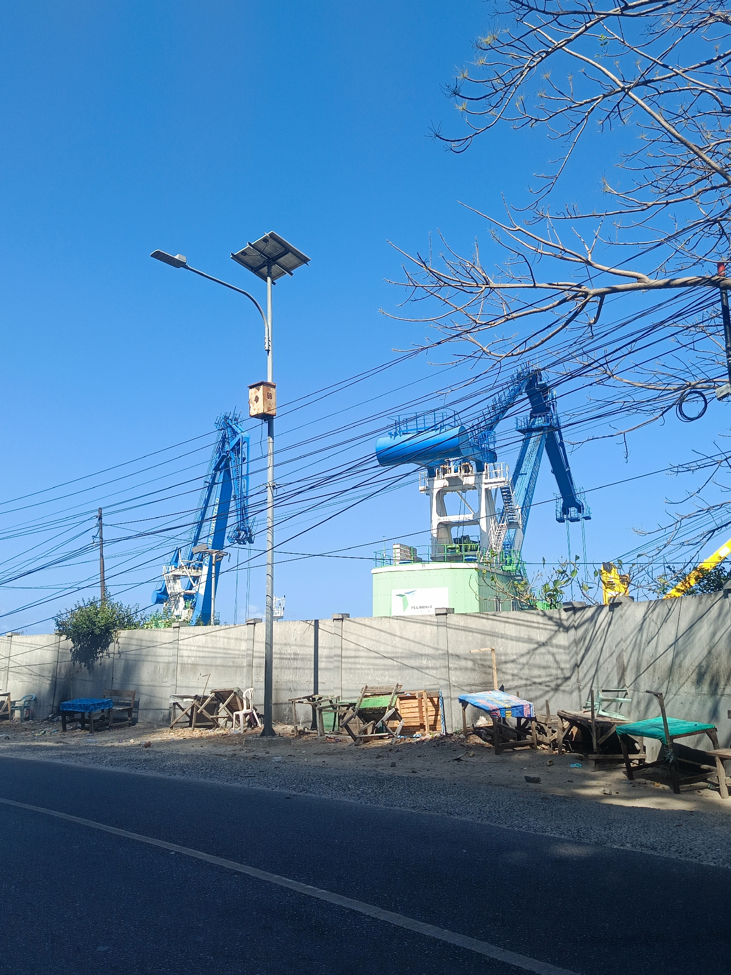 The image captures a bright, sunny day in Gorontalo, Indonesia, with a clear blue sky dominating the upper portion. In the background, two large blue cranes are visible, suggesting a port or construction area. One crane is positioned to the left, and another, larger one, is in the center. These cranes are part of a facility with a light green building beneath them, marked with a white logo that includes the text "PELINDO I". A concrete wall runs horizontally across the middle ground, separating the road from the cranes and building.
In the foreground, a paved road with a white line marking its edge runs from the bottom left to the bottom right of the frame. Along the side of the road, adjacent to the concrete wall, there are several makeshift stalls or tables constructed from wood and covered with various colored tarps, including blue and green. Some of these structures appear weathered and possibly abandoned. Debris is scattered on the ground around these stalls.
To the left of the frame, a tall lamppost stands prominently, equipped with a solar panel and a small rectangular box attached to its pole, possibly an electrical enclosure or a birdhouse, with the number "69" visible on it. Overhead, a network of thick electrical wires crisscrosses the scene, partially obscuring the view of the cranes and the sky. Bare branches of a tree extend into the frame from the upper right, their stark silhouettes contrasting with the bright sky. The overall scene depicts an industrial or port area in Gorontalo, with elements of infrastructure, commerce (represented by the stalls), and the natural environment.