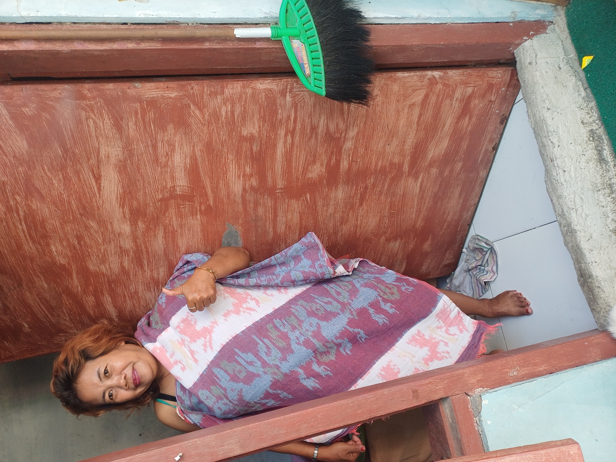 In Pulungan, Indonesia, a woman with reddish-brown hair stands in a doorway, wrapped in a patterned cloth. She is offering a thumbs-up gesture with her right hand. A green broom with black bristles is leaning against the right side of the doorframe, near a rusty brown door. The floor is tiled, and a light blue painted wall is visible to the left of the doorway. Wooden elements frame the entrance, and a concrete threshold is present.