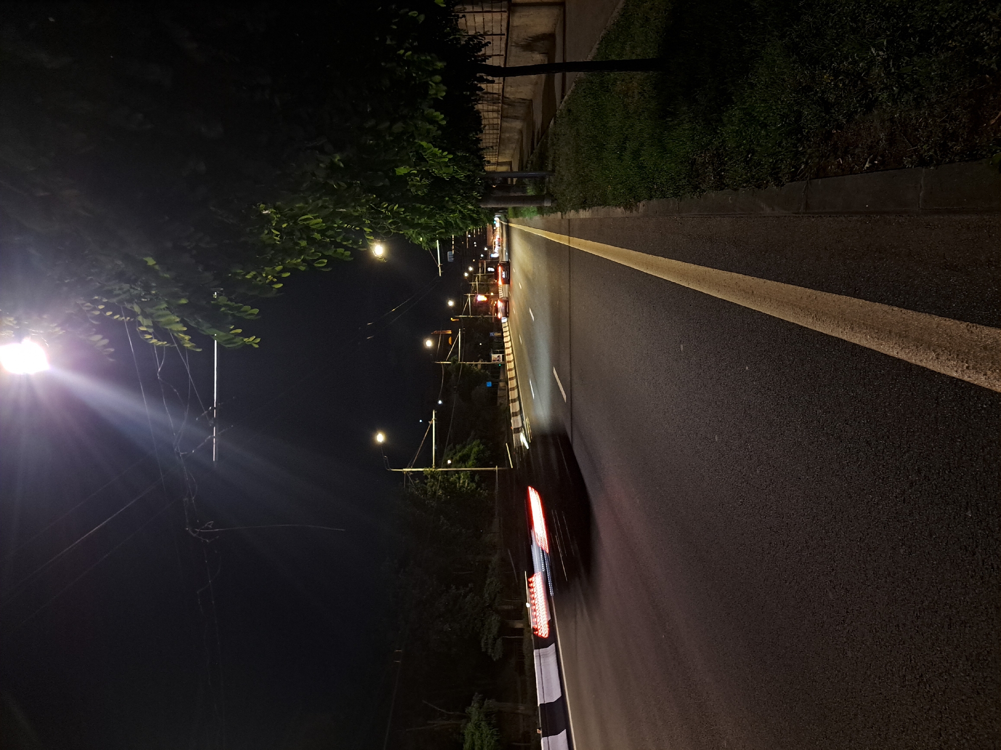 Captured at night in Yerevan, Armenia, the image presents a street scene illuminated by streetlights. The dark asphalt road features a solid yellow line marking the lane division. A car is captured in motion, its blurred taillights indicating movement. Vegetation lines the edge of the road, transitioning into a sidewalk or curb. The setting is urban with visible electrical lines intersecting the sky, and the lighting conditions cast a stark contrast.