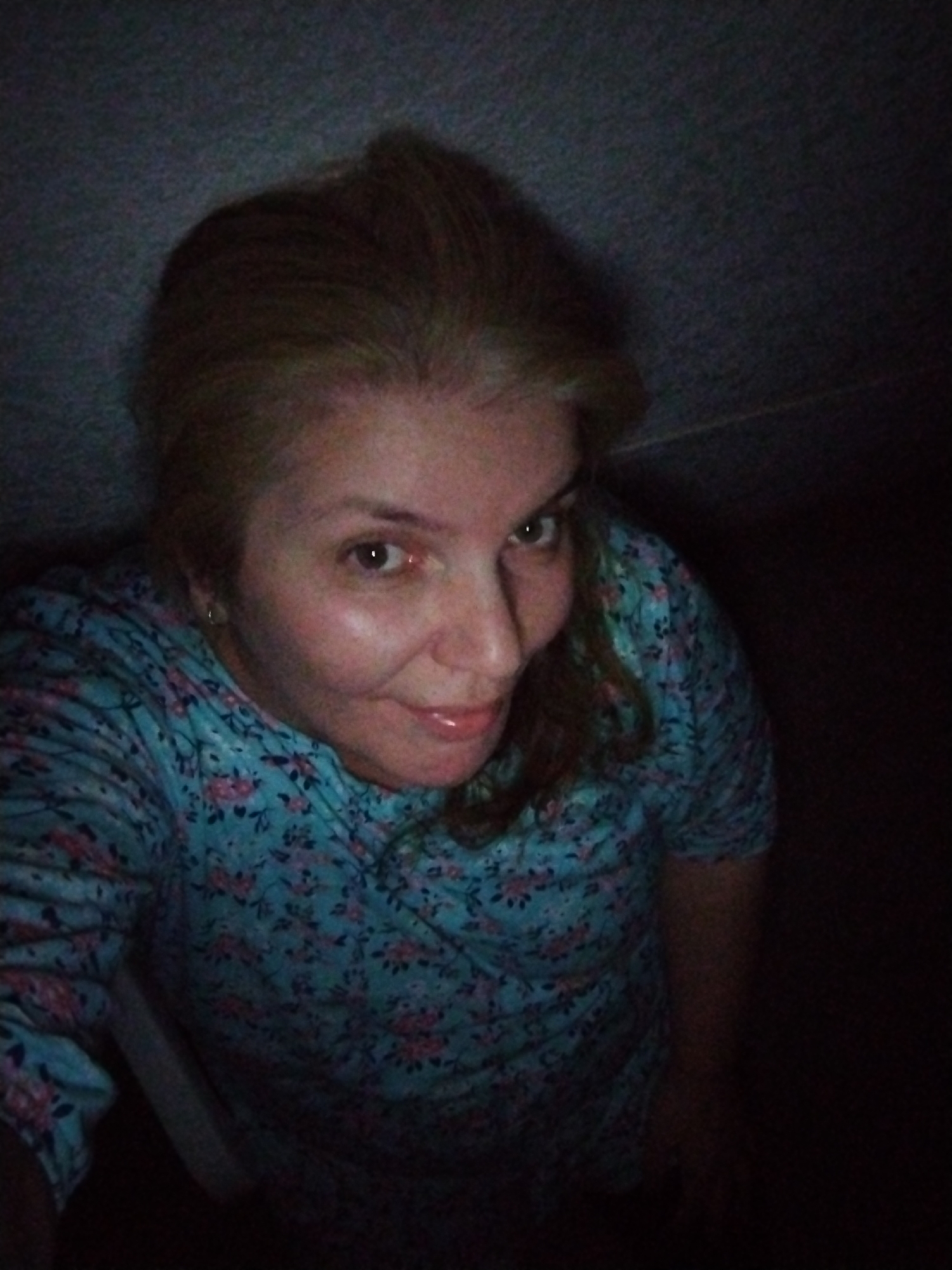 A woman is posing for a selfie in what appears to be a dimly lit room. She has fair skin, light brown hair, and is wearing a blue shirt with a floral pattern. The background is a dark, textured wall, possibly in a residential setting within Al Mas'oudi, United Arab Emirates. The lighting suggests the photo was taken indoors, possibly at night or in an area with low ambient light.