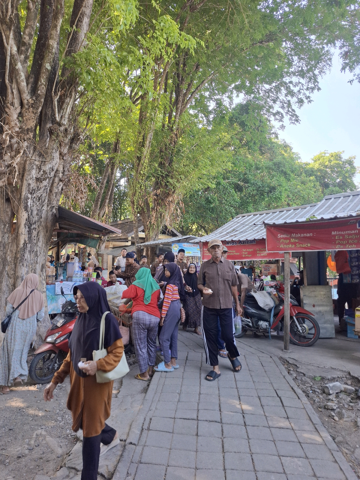 The image depicts a bustling street scene in Ngawi, Indonesia, under a clear sky and dappled sunlight filtering through overhead trees. The foreground shows a paved walkway with a few individuals walking. A woman in a brown abaya and black hijab walks from left to right, carrying a light-colored tote bag. Behind her, a man in a brown shirt and dark pants with white stripes walks in the same direction. To the left, a woman in a light blue patterned dress and beige hijab is visible.
The middle ground is lined with several food stalls and small shops, indicated by awnings and signage. One stall has a red banner that reads "Sedia Makanan: - Pop Mie - Aneka Snack" and "Minuman: - Es Salju - Pop Ice - Es Teh". Another stall has "WARUNG KECIL" visible. Several motorcycles, including a red one, are parked near the stalls. People are gathered around the stalls, some appear to be purchasing food and drinks, while others are conversing. A water dispenser is visible at one of the stalls.
The background is dominated by large trees with dense green foliage, suggesting a natural or park-like setting within the city. The overall atmosphere appears to be that of a local market or a lively street corner with commercial activity.