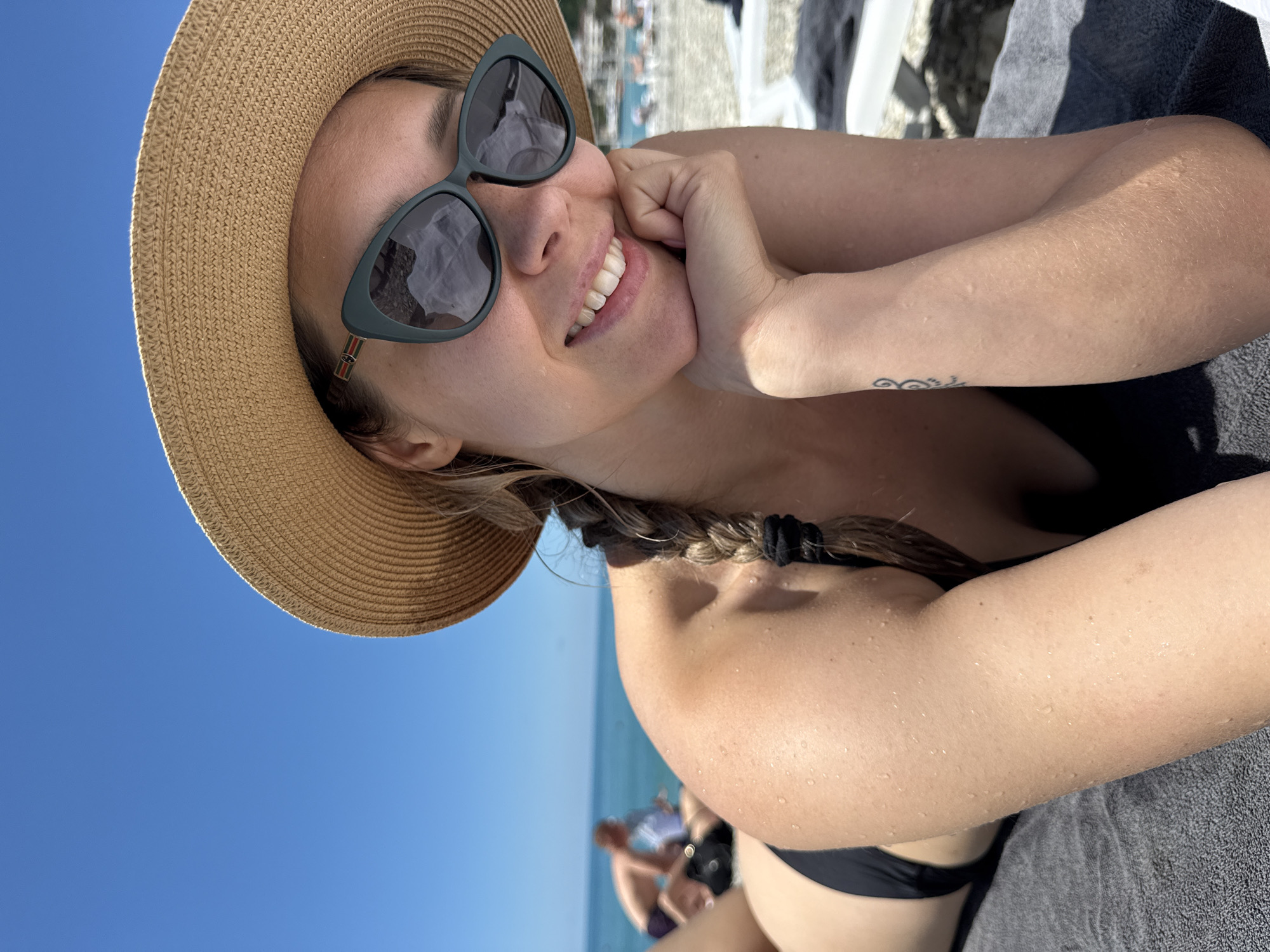 A woman wearing a black bikini and a straw hat with sunglasses lies on a beach towel on a sunny day. Her hand supports her chin as she smiles at the camera. Water droplets are visible on her skin. In the background, the blue sea and a beach with other people can be seen. The scene is set at a beach resort in Nebug, Russia.