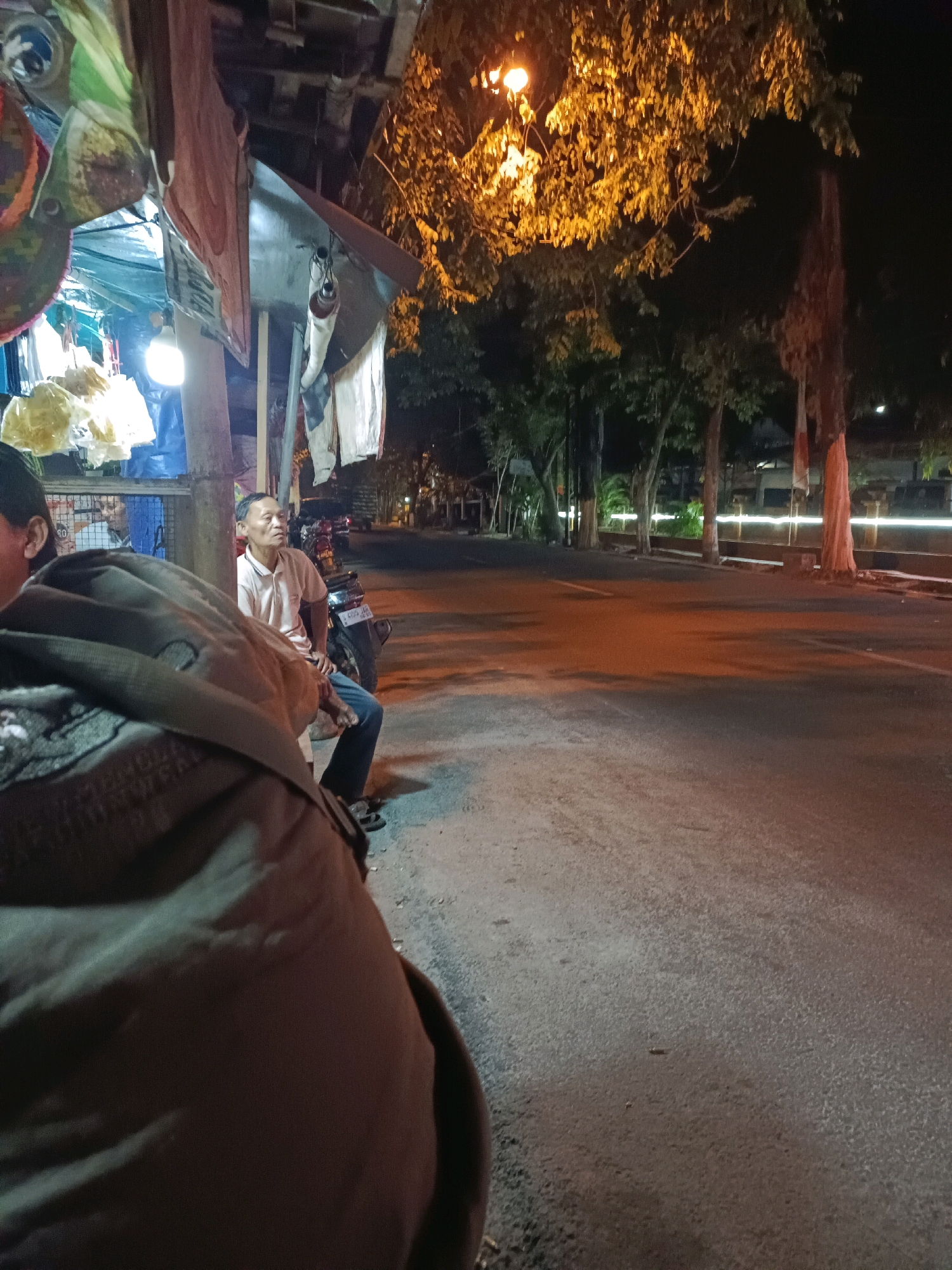 The image captures a nighttime scene at a roadside stall in Lamongan, Indonesia. In the foreground, a person is partially visible from the back, wearing a dark jacket. To their right, a man in a light-colored polo shirt and jeans is seated on a motorcycle, looking to the side. Behind the man, the stall displays various items, including what appear to be bags of snacks or goods, covered by tarps and fabric. A bright light illuminates the stall area.
The background shows a road with traffic lights and trees lining the sidewalk. The street appears wet in places, reflecting the warm glow of streetlights and the stall's illumination. The overall atmosphere suggests a quiet, late-night street scene in a moderately urban environment.