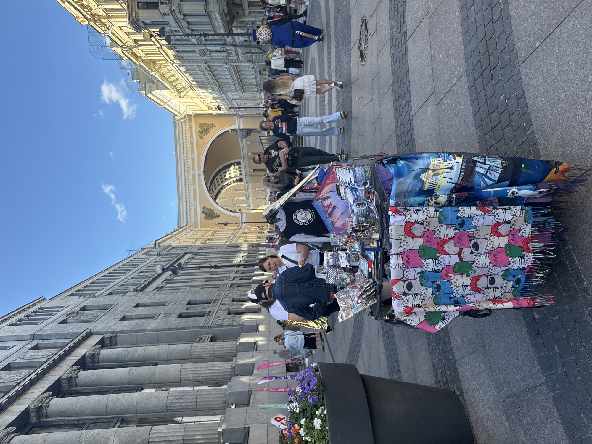 In Saint Petersburg, Russia, on a sunny day, people walk along a wide, cobblestone pedestrian street lined with classical architecture. To the left, a large building with imposing columns dominates the view, while to the right, a row of ornate, yellow-painted buildings stretches into the distance, culminating in a grand archway. In the foreground, a street vendor's table displays a variety of souvenirs, including colorful scarves patterned with cats, mugs, and other trinkets. Several people are browsing the merchandise, and others are walking by. A person in a leopard costume walks past the stall, drawing attention. The scene captures a lively atmosphere in a historic urban setting.