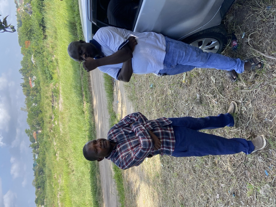 Two men stand outdoors near a dirt road. One leans against a silver car, wearing a white collared shirt, blue jeans, and sandals. The other stands with arms crossed, dressed in a maroon plaid shirt and blue pants with brown shoes. Grass and trees form the background, with scattered buildings visible in the distance. The sky is partly cloudy.