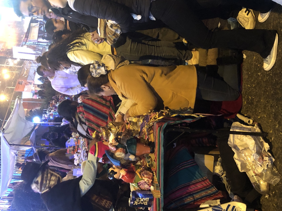 A bustling outdoor market scene unfolds, illuminated by warm lighting. People of varying ages and attire browse a vendor's table laden with colorful goods. Handmade crafts, trinkets, and textiles are on display, appealing to customers with their vibrant patterns and designs. A white tent can be seen in the background with many other people walking around. The atmosphere suggests a lively evening of shopping and cultural exploration.