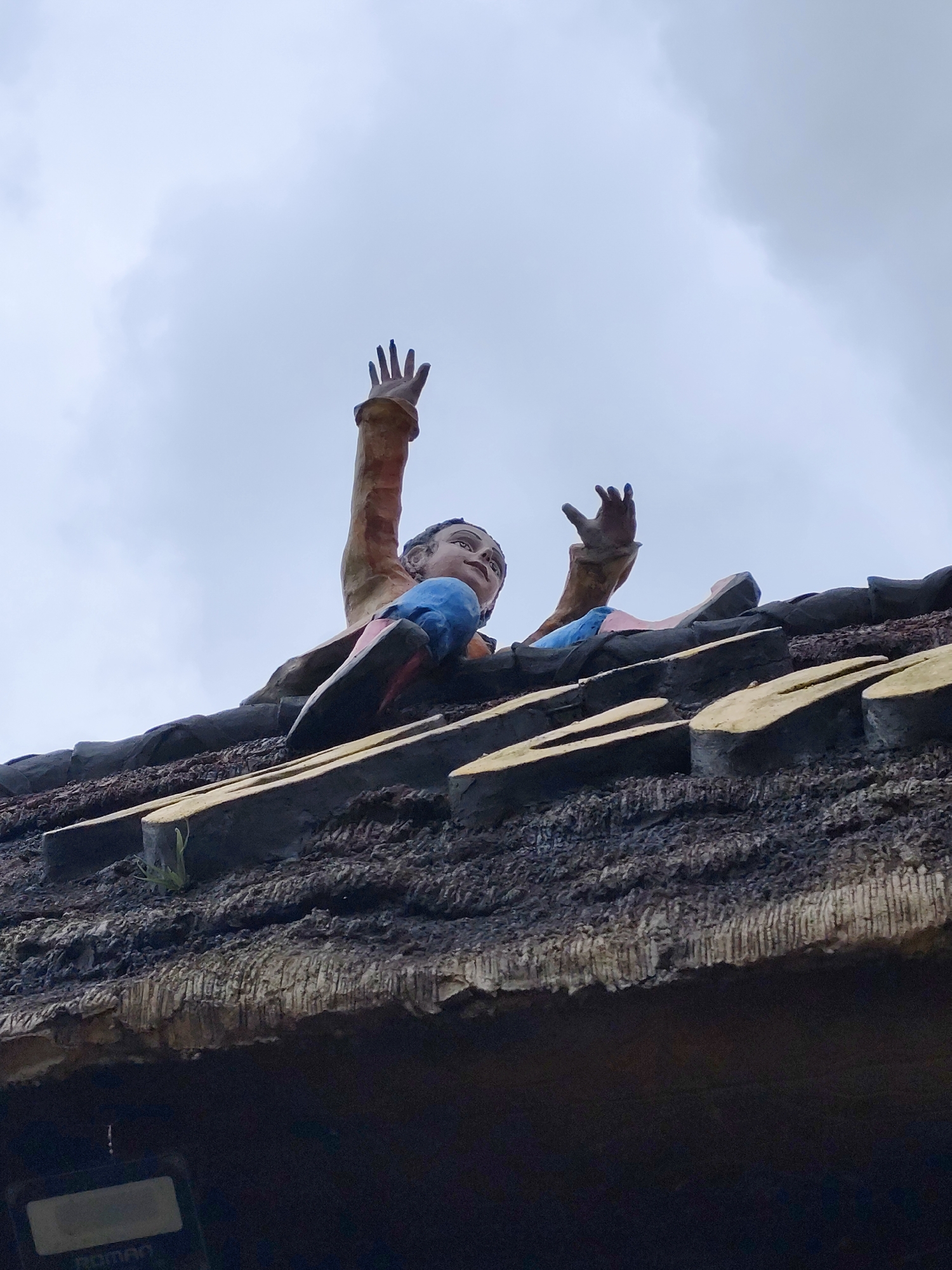 A striking low-angle photograph captures a vibrant, humanoid statue appearing to emerge dramatically from a dark, textured roof against a bright, overcast sky. The figure, depicted from the waist up, is positioned with both arms raised and hands open, suggesting a gesture of struggle, reaching, or calling for help. Its face, looking slightly upwards and to the right, has an open mouth conveying an expression of distress or intense effort. The statue is adorned in a light brown long-sleeved top and blue lower garments with hints of red.
The roof is thick and heavily textured, resembling a traditional thatched roof or one crafted from dark, organic materials. Several lighter, yellowish, plank-like elements are horizontally integrated into its surface. The sky overhead is uniformly bright and cloudy, indicative of an overcast daytime. Partially visible in the bottom-left corner, beneath the eaves of the roof, is a dark electronic device with a rectangular screen displaying the word "ROMAN".
