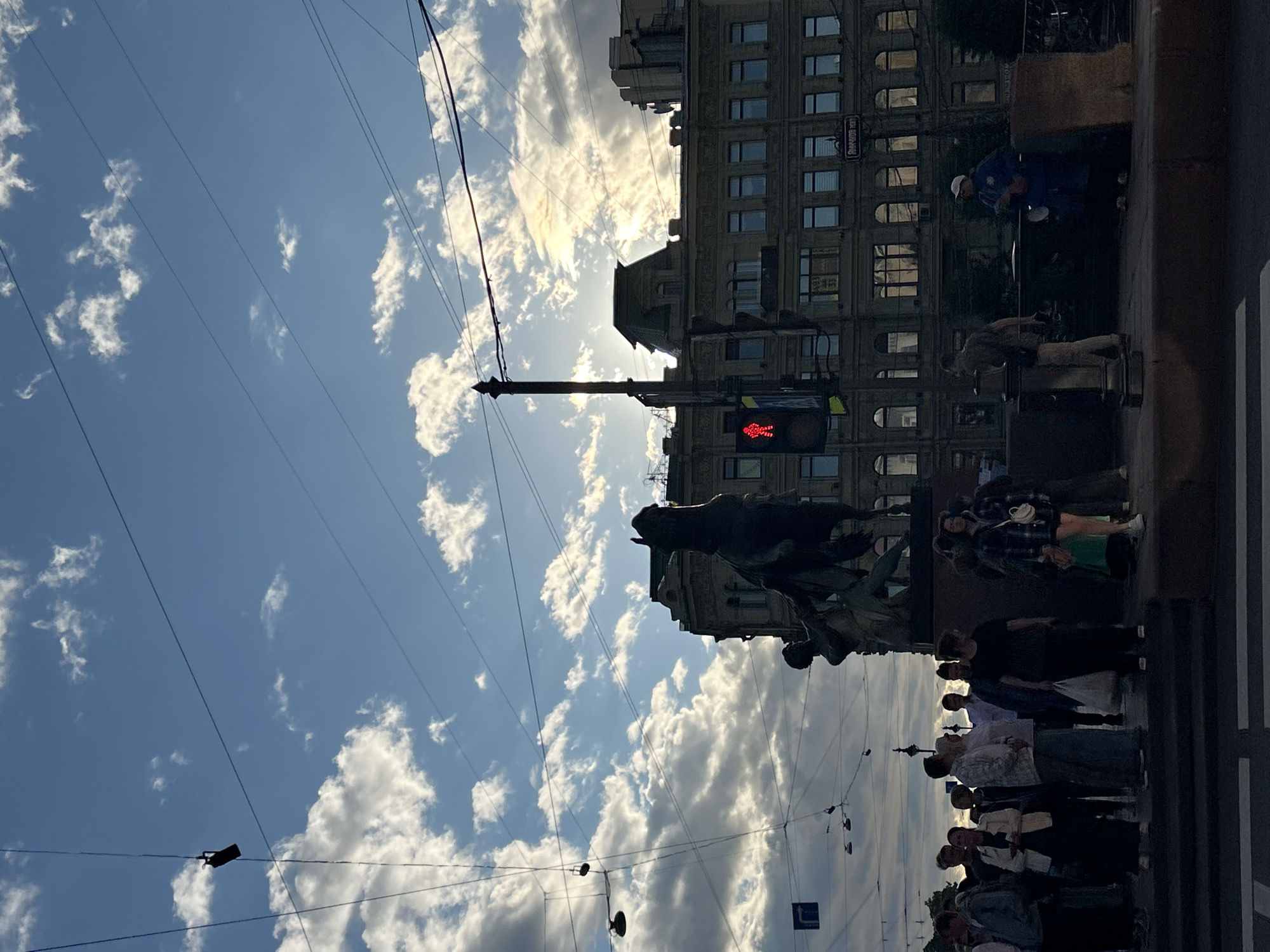 The image captures a street scene with a statue of a horse and rider in the foreground, silhouetted against a partly cloudy sky. A group of people are gathered on a raised platform or sidewalk next to the statue, some looking towards the camera, others interacting with each other. A traffic light displaying a red pedestrian signal is visible to the right of the statue, adjacent to a multi-story building. Overhead, a network of wires and streetlights crisscrosses the sky. The building behind the statue appears to be an older, ornate structure with numerous windows. The overall atmosphere suggests an urban environment during daylight hours, with the sun partially obscured by clouds, casting a dramatic light on the scene.