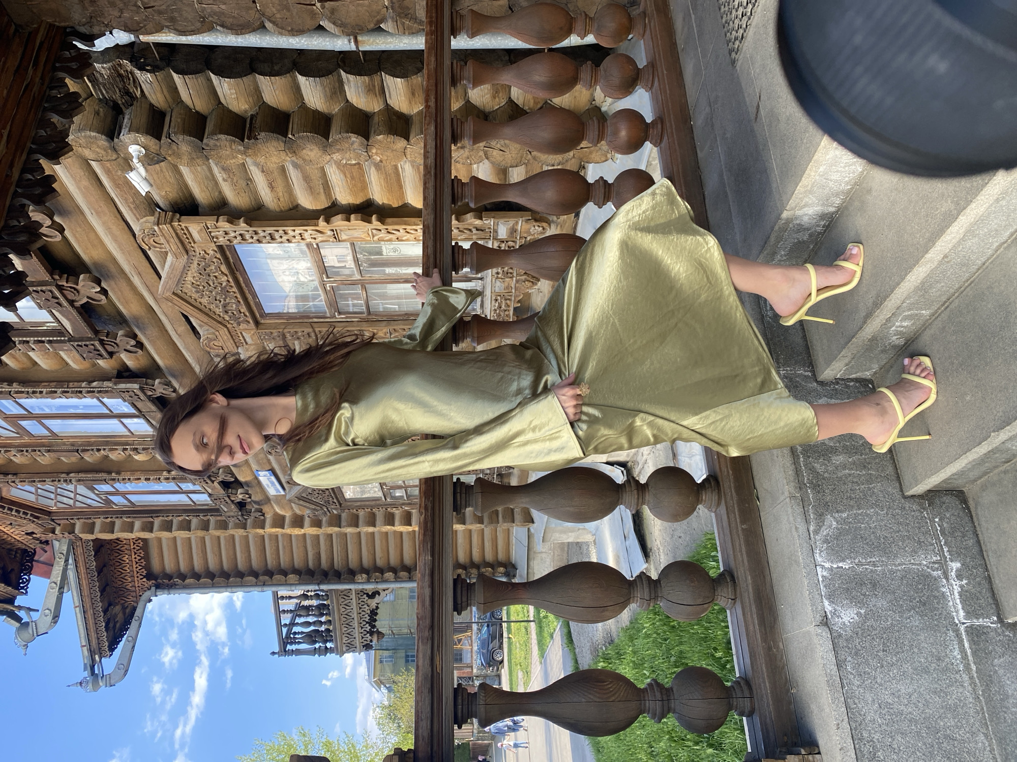 A woman in a long, green dress and yellow high-heeled sandals is posing on the porch of the Sevastyanov House in Yekaterinburg, Russia. She is standing next to a wooden railing, and the building's ornate wooden facade is visible in the background. The sky is blue with some clouds. The porch is made of stone, and there is grass and a sidewalk visible in the background. People are walking on the sidewalk in the background.
